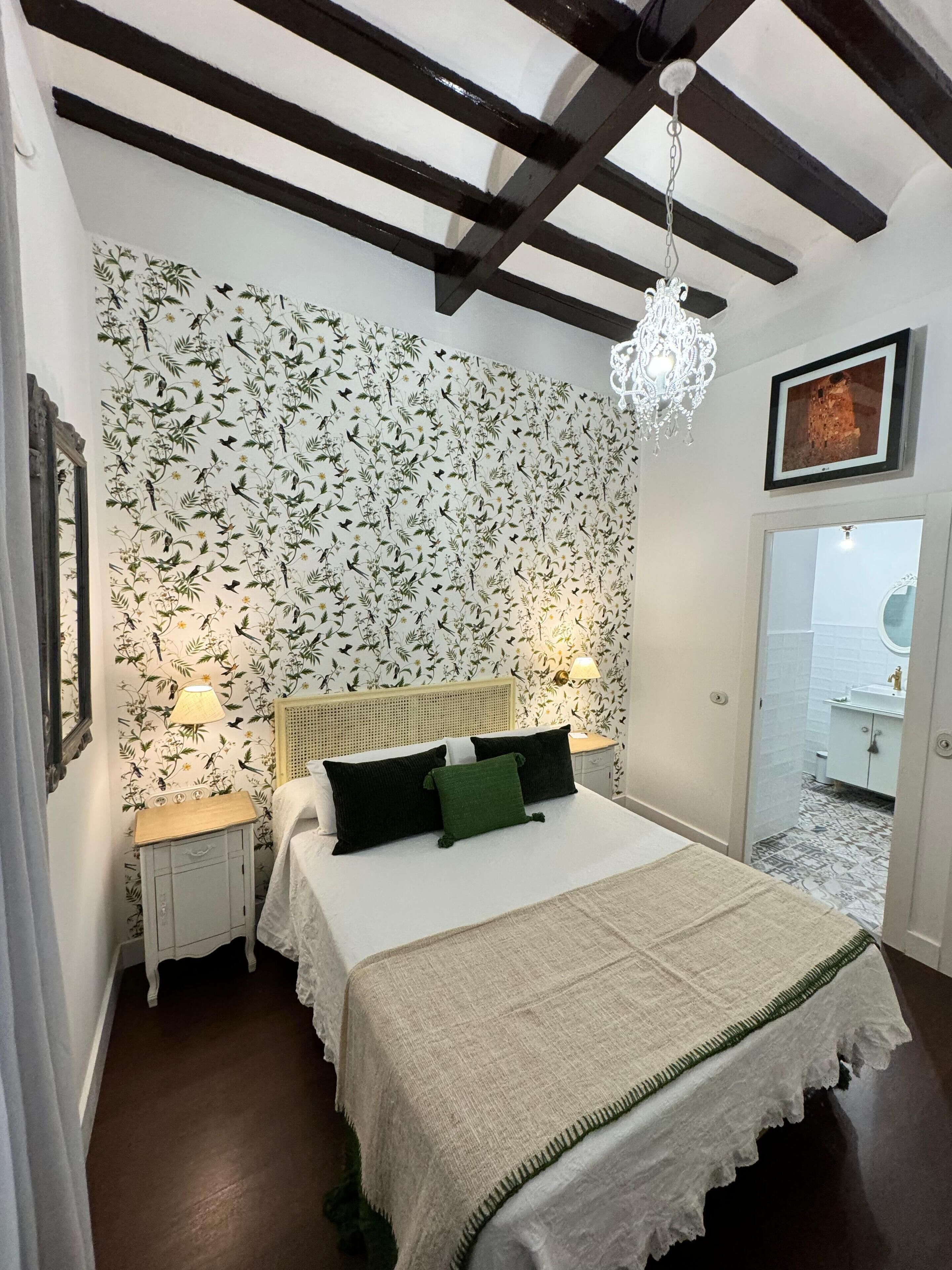 Un dormitorio con una cama, dos mesitas de noche, una araña, una pared cubierta de papel tapiz floral y una puerta que da al baño.