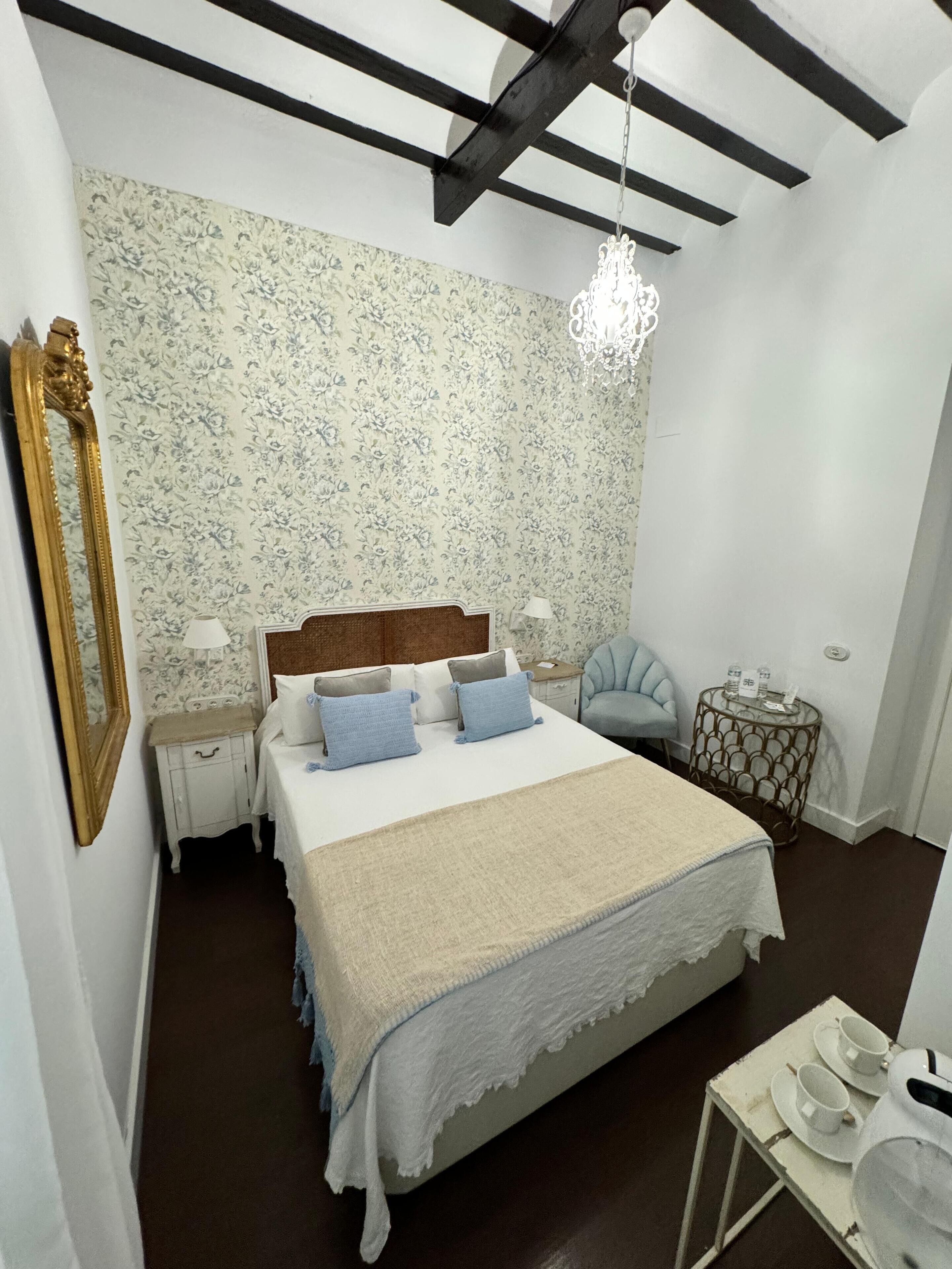 Un dormitorio con una cama, mesitas de noche, una silla, una pequeña mesa con tazas y un plato, y una araña.