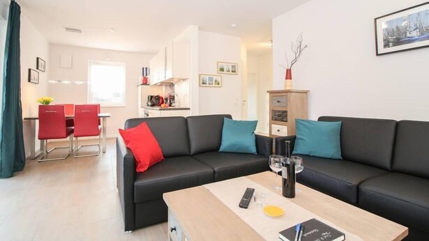 Un salon moderne avec un canapé noir, des coussins rouges et turquoise, une table basse en bois sur laquelle se trouvent une télécommande, un verre et un livre.