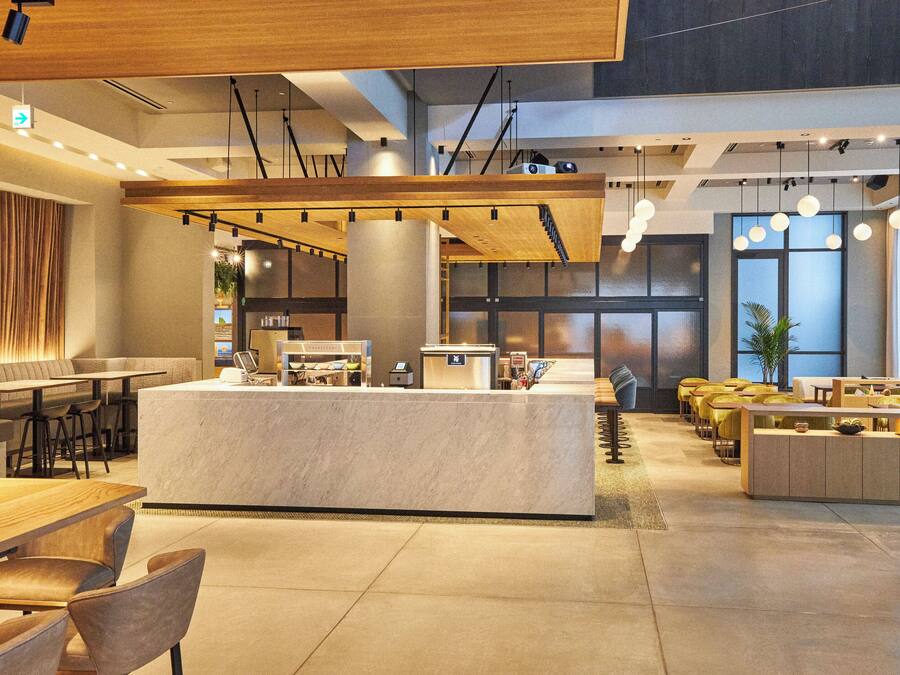 A modern hotel lobby with a central reception desk, seating areas, and a large window providing natural light.