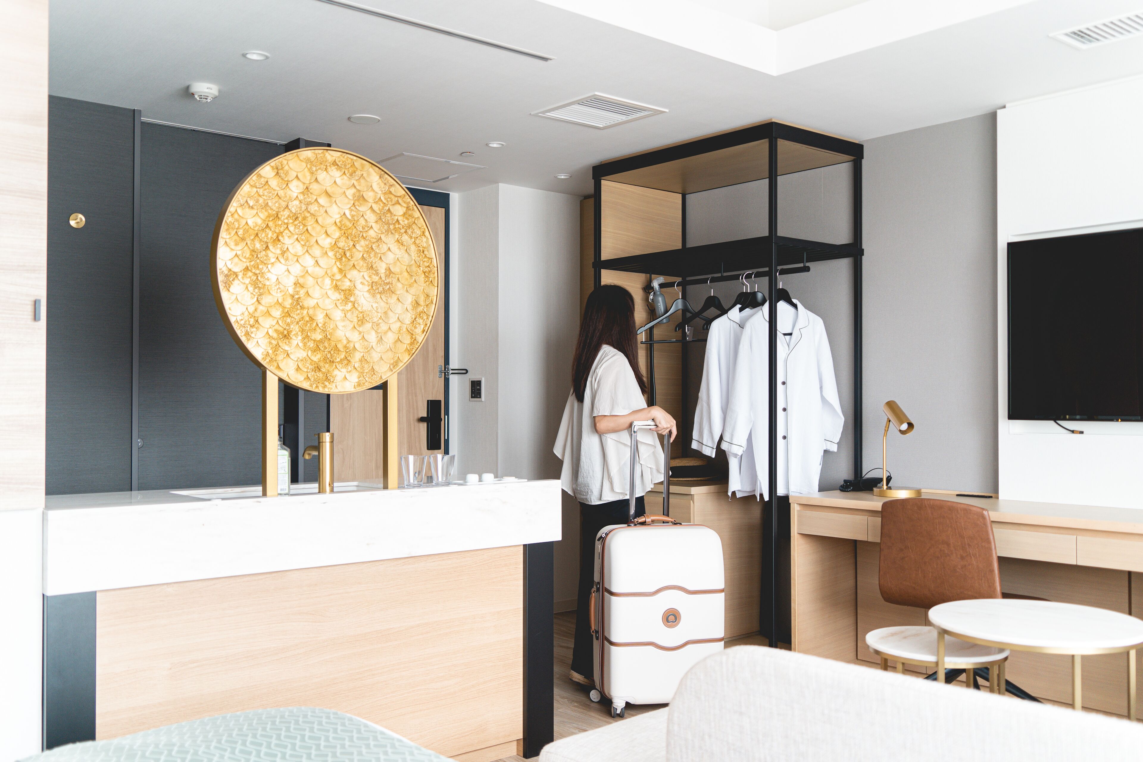 A modern hotel room with a large round mirror, a wardrobe with a black frame, a desk with a chair, and a small table.