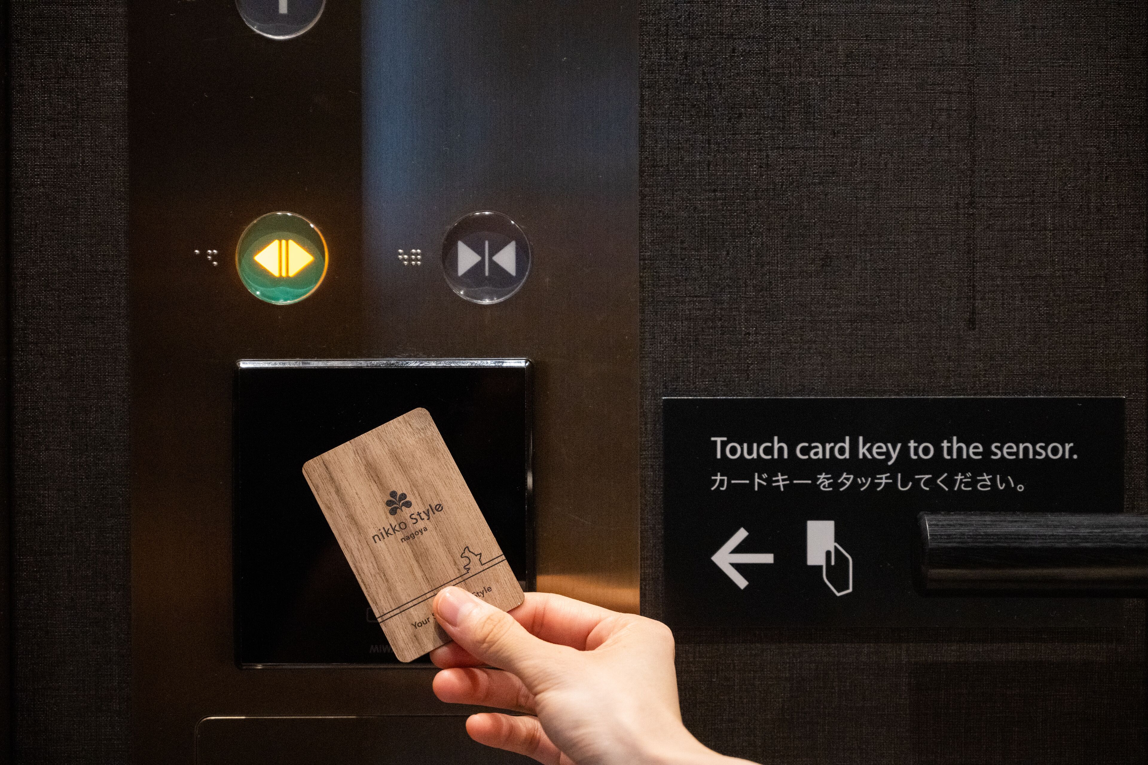 A hand holding a wooden card with "nikko style" written on it, placed in an elevator with a sign instructing to "Touch card key to the sensor."