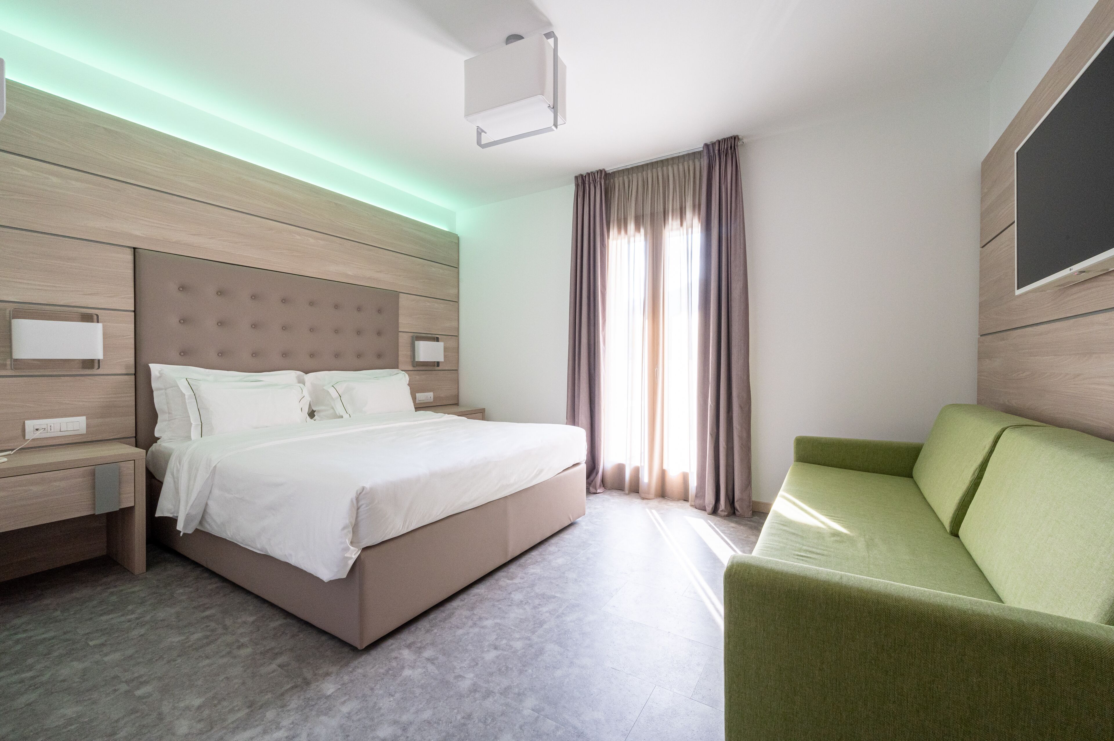 A modern hotel room with a large bed, a green sofa, and a flat-screen TV on the wall.