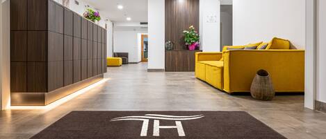 A hotel reception area with a dark brown carpet with "TRILAGO HOTEL" written on it, a wooden reception desk, and a yellow sofa with cushions.