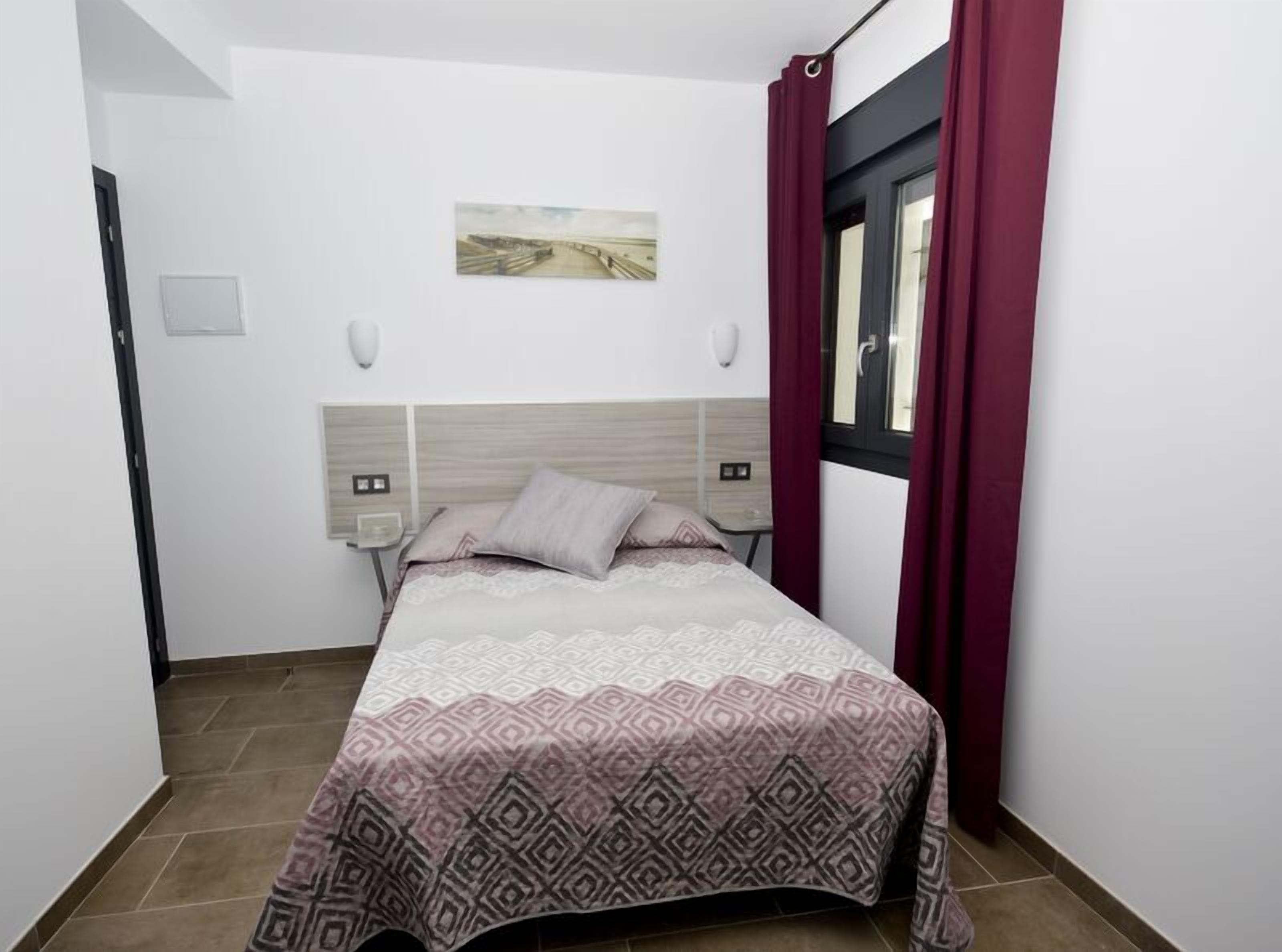 A bedroom with a bed, a nightstand, a wall-mounted lamp, a window with curtains, and a painting on the wall.