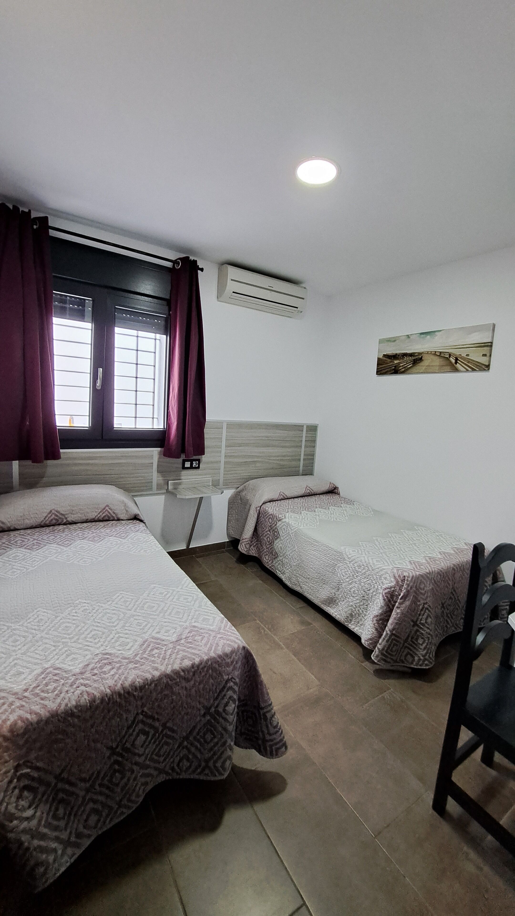 Una habitación con dos camas, una ventana con cortinas, un equipo de aire acondicionado y un cuadro en la pared.