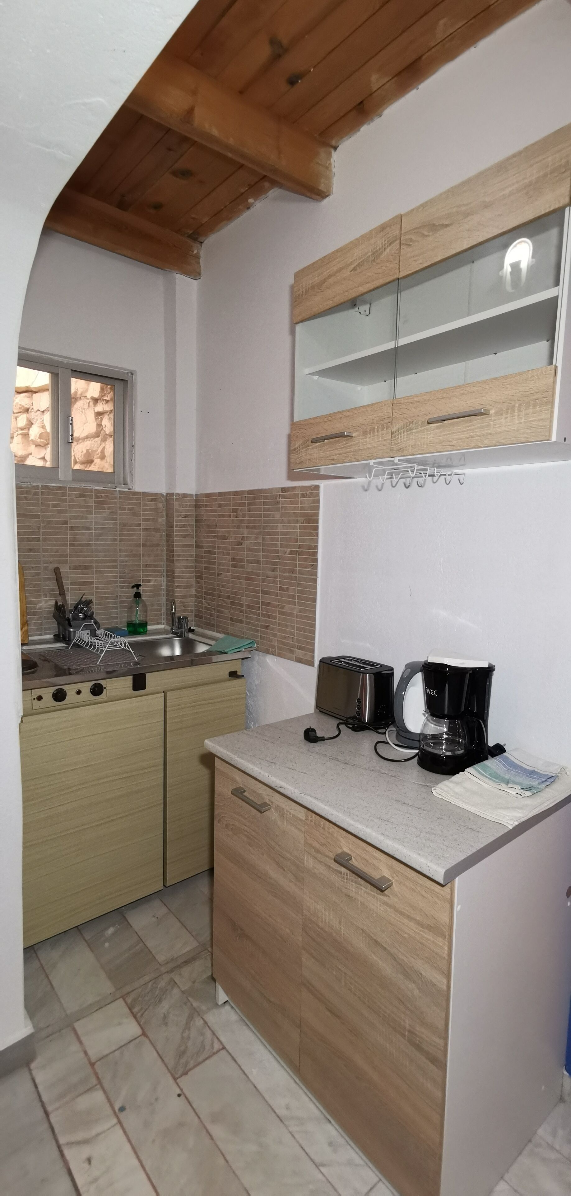 Cuina compacta amb sostre de fusta, una aixeta de sobretaula, torradora, cafetera i finestra amb vista a una paret de pedra.