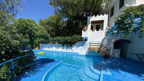 Una piscina con agua azul cristalina, rodeada de árboles y un edificio blanco con una escalera de madera.