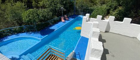 Una piscina con una valla decorada con motivos de castillo, rodeada de abundante vegetación y una terraza de madera.