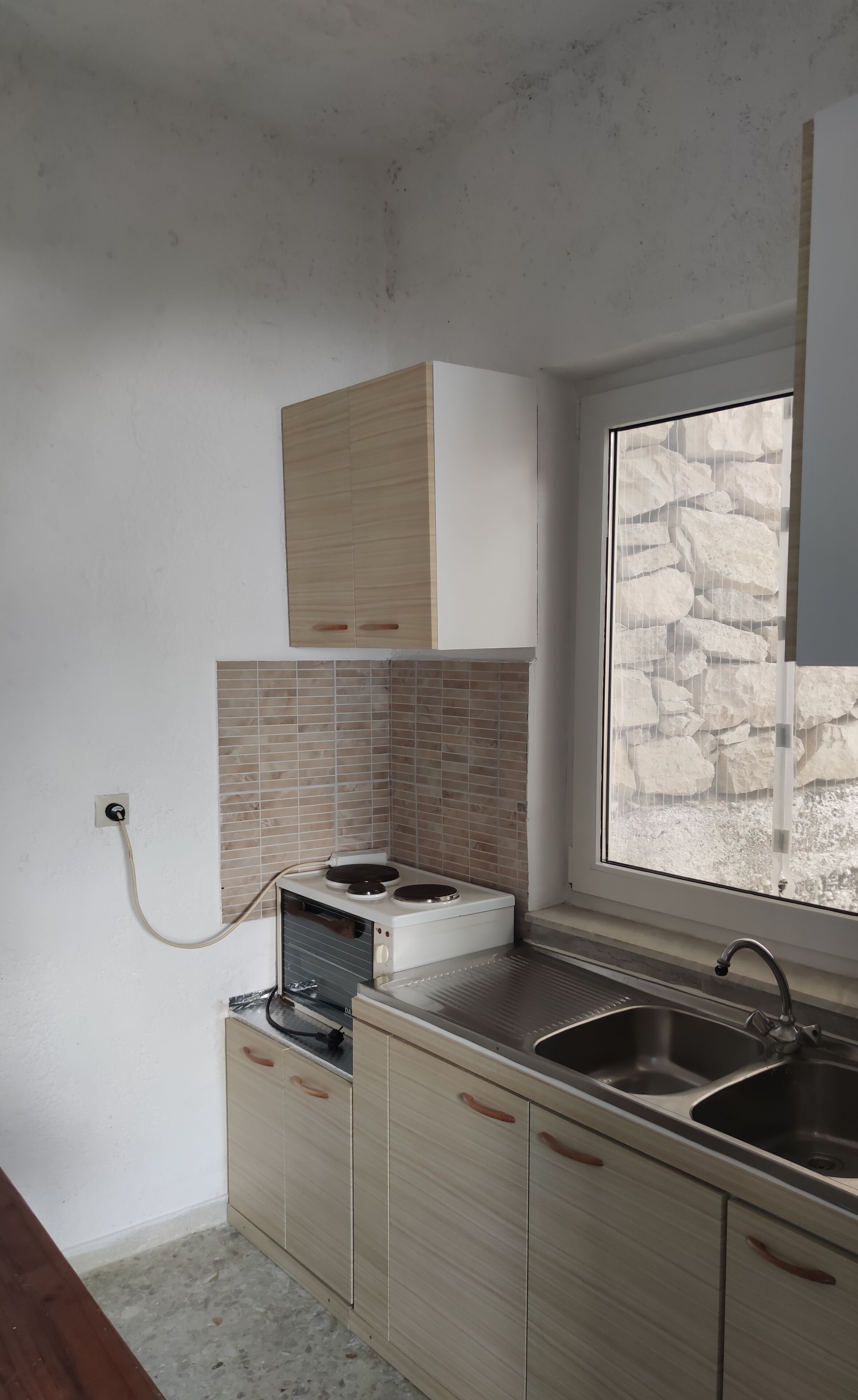 A kitchen with a built-in stove, a double sink, wooden cabinets, and a stone wall visible through a window.