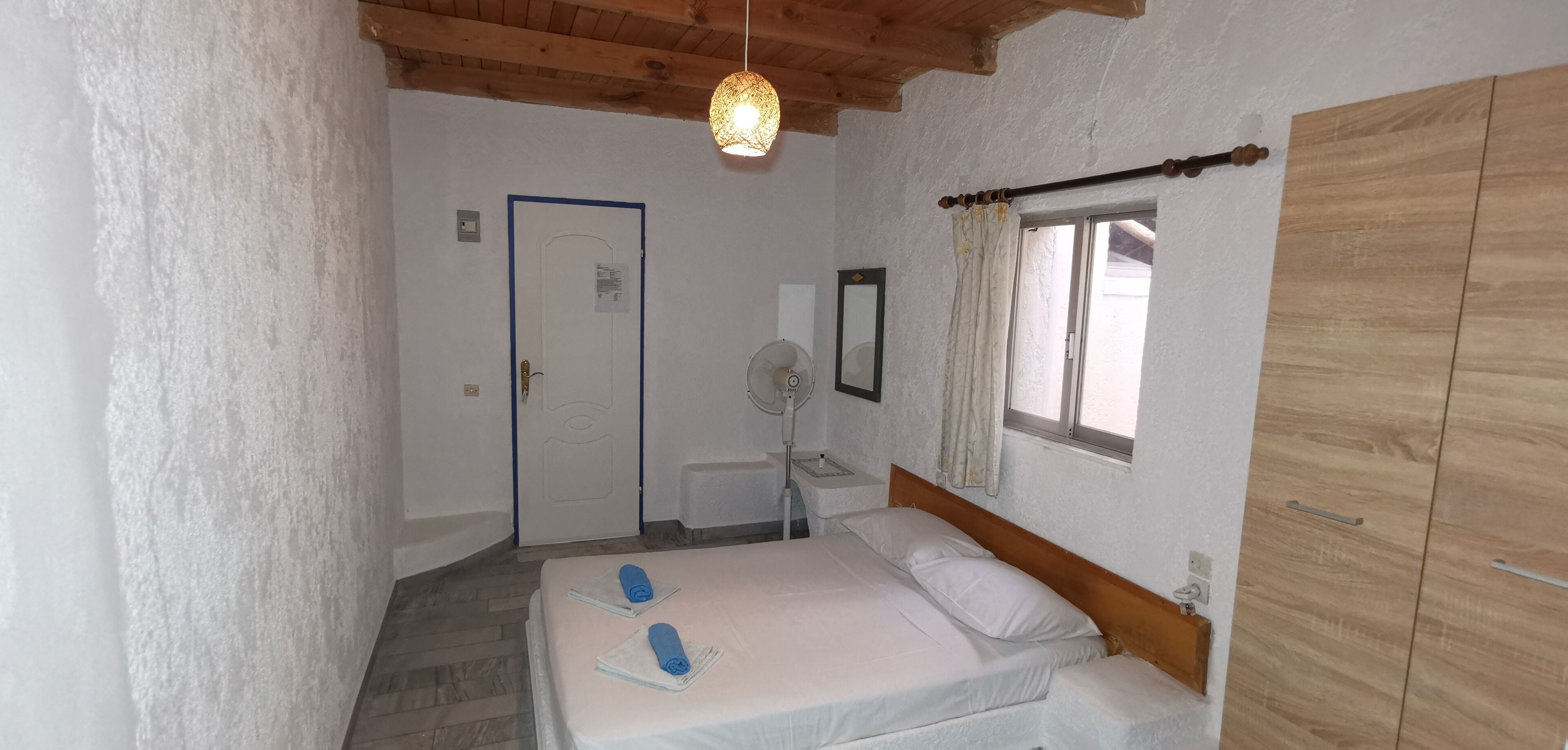 A bedroom with a bed, a bedside table, a mirror, a window with curtains, and a wooden wardrobe.