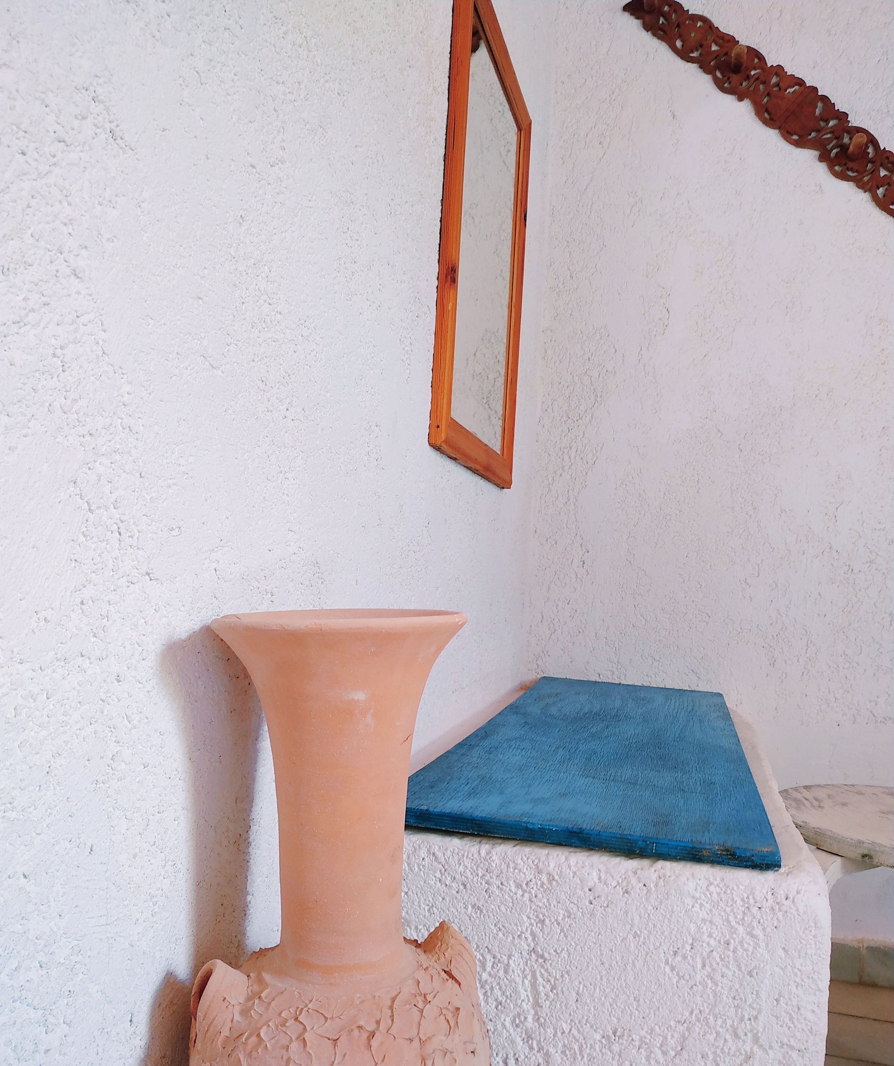 A terracotta vase on a white textured wall, a blue wooden board, and a wooden-framed mirror.