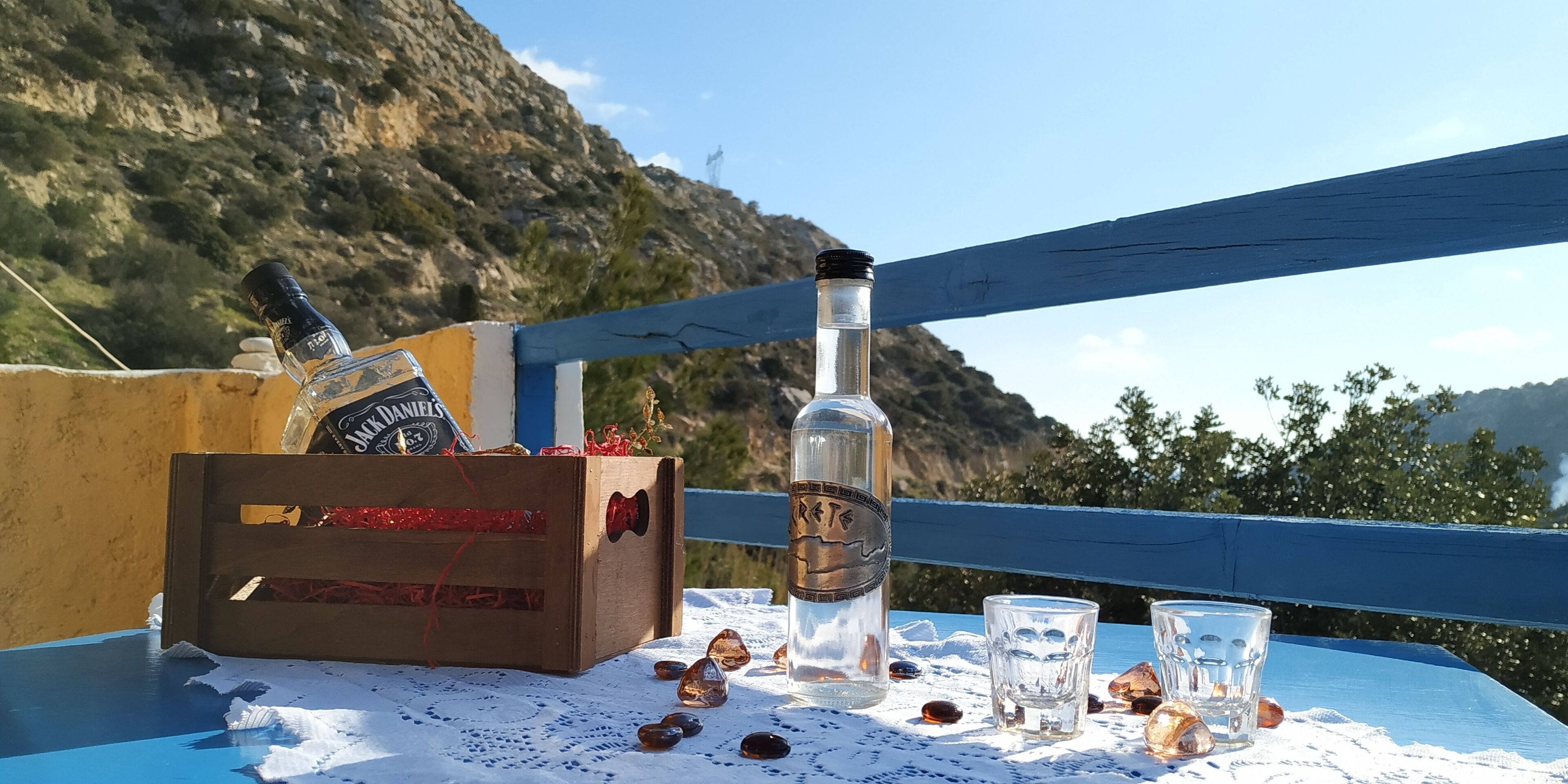 A wooden crate with a bottle of Jack Daniel's whiskey and a bottle of water on a table with a lace tablecloth, surrounded by scattered amber-colored stones, on a balcony with a blue railing and a mountainous backdrop.