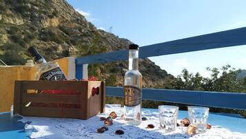 A wooden crate with a bottle of Jack Daniel's whiskey and a bottle of water on a table with a lace tablecloth, surrounded by scattered amber-colored stones, on a balcony with a blue railing and a mountainous backdrop.