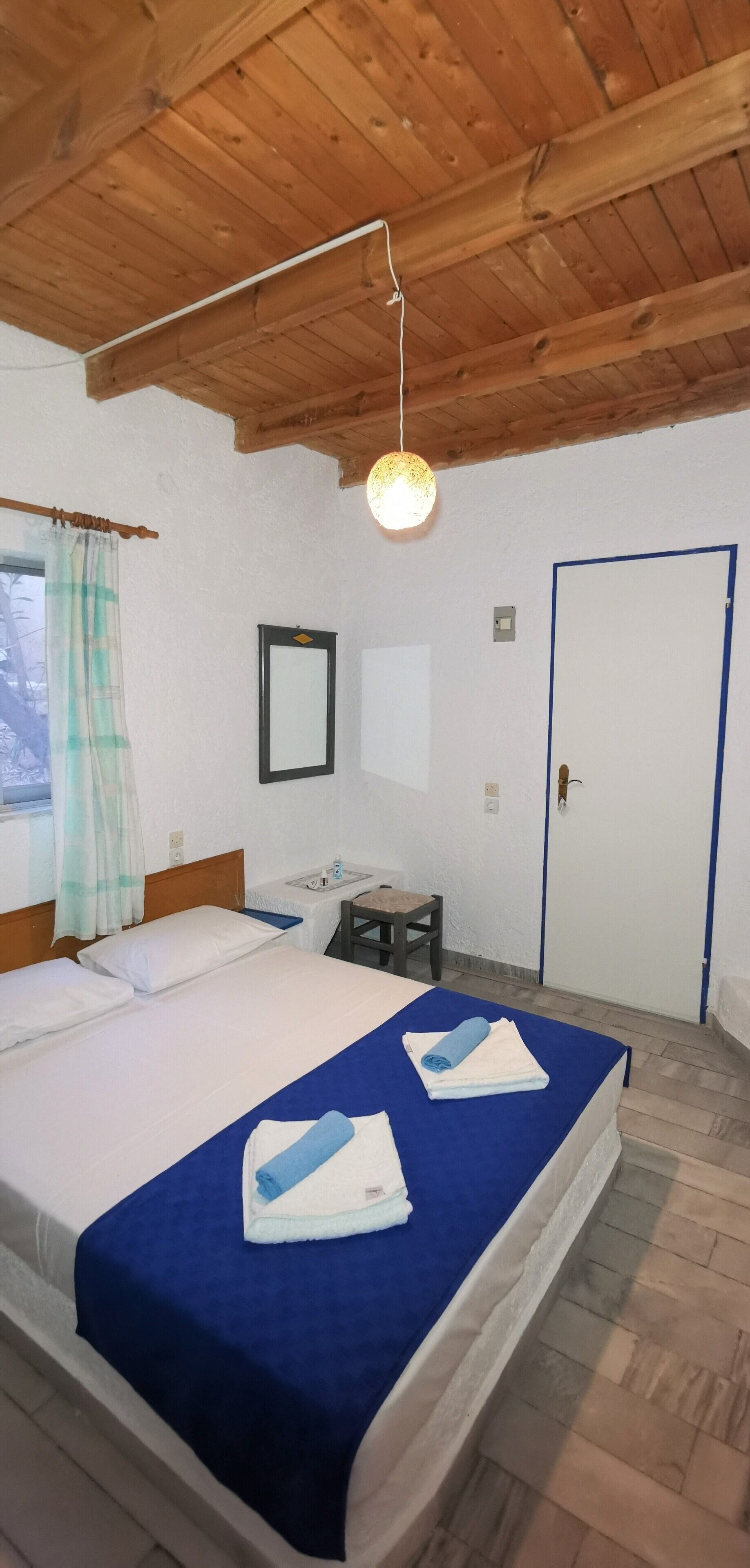 A room with a wooden ceiling, a bed with blue bedding, a bedside table, a chair, a small table with a lamp, and a door.