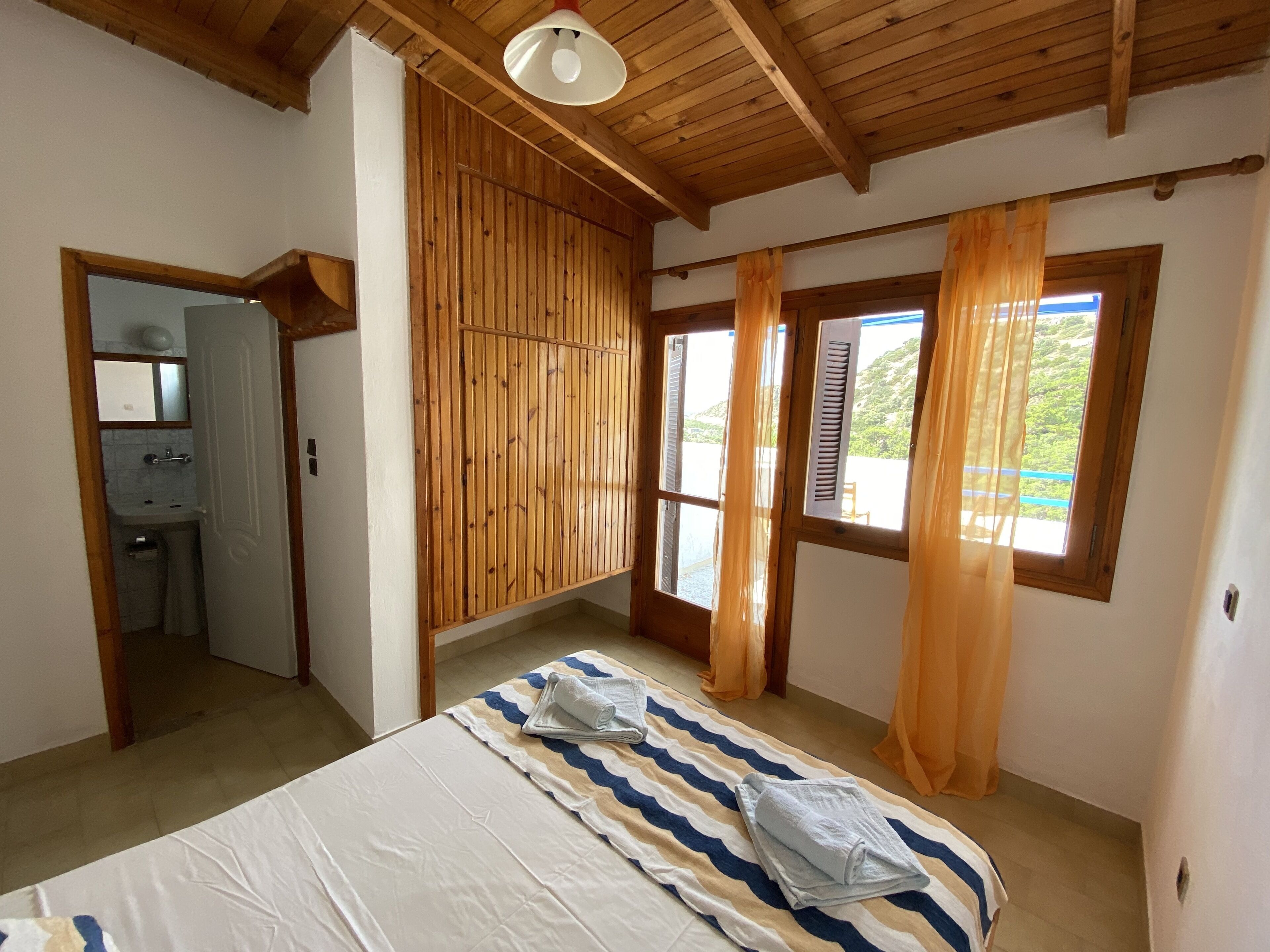 Une chambre à coucher avec un lit, une armoire en bois, une salle de bain et une fenêtre avec des rideaux.