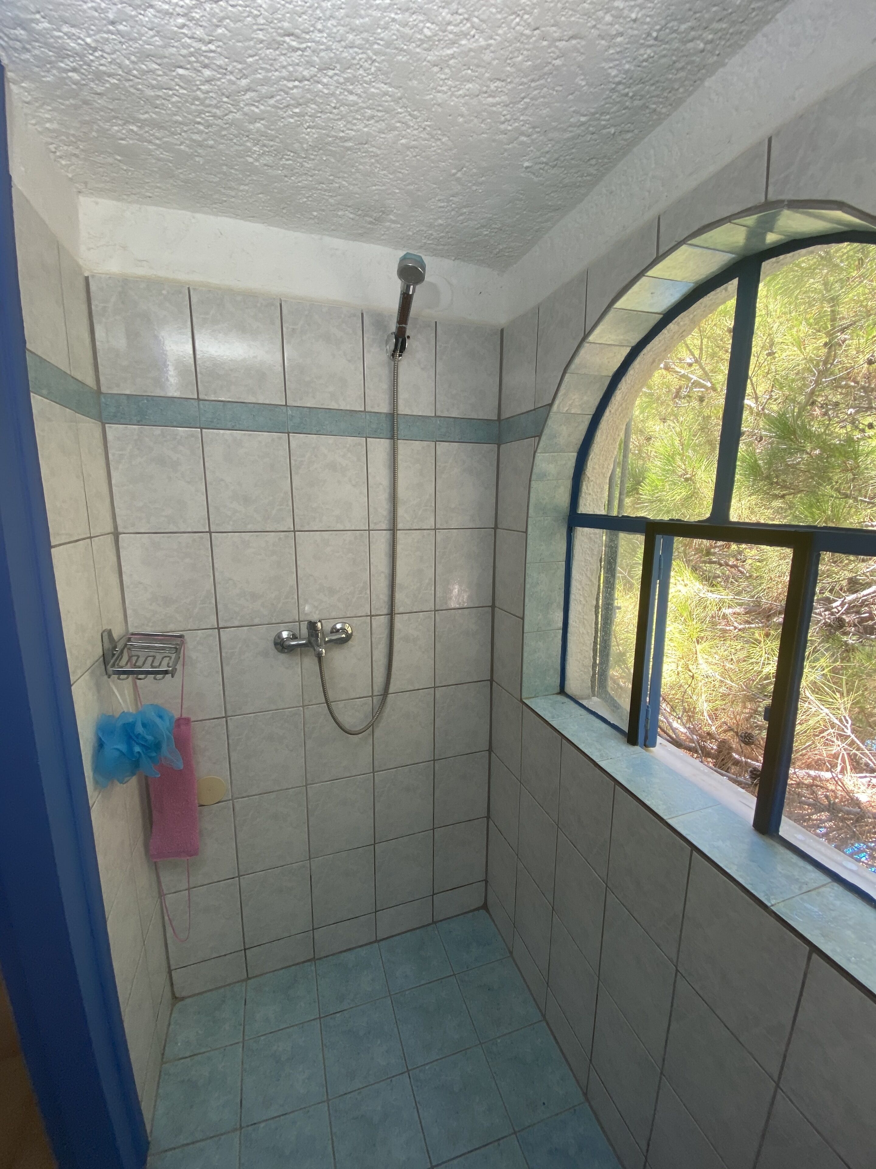 Une petite salle de douche carrelée, avec une fenêtre et un étagère en métal.