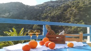 Una mesa con naranjas y dos copas de vino sobre un mantel azul, con vistas a un paisaje montañoso.