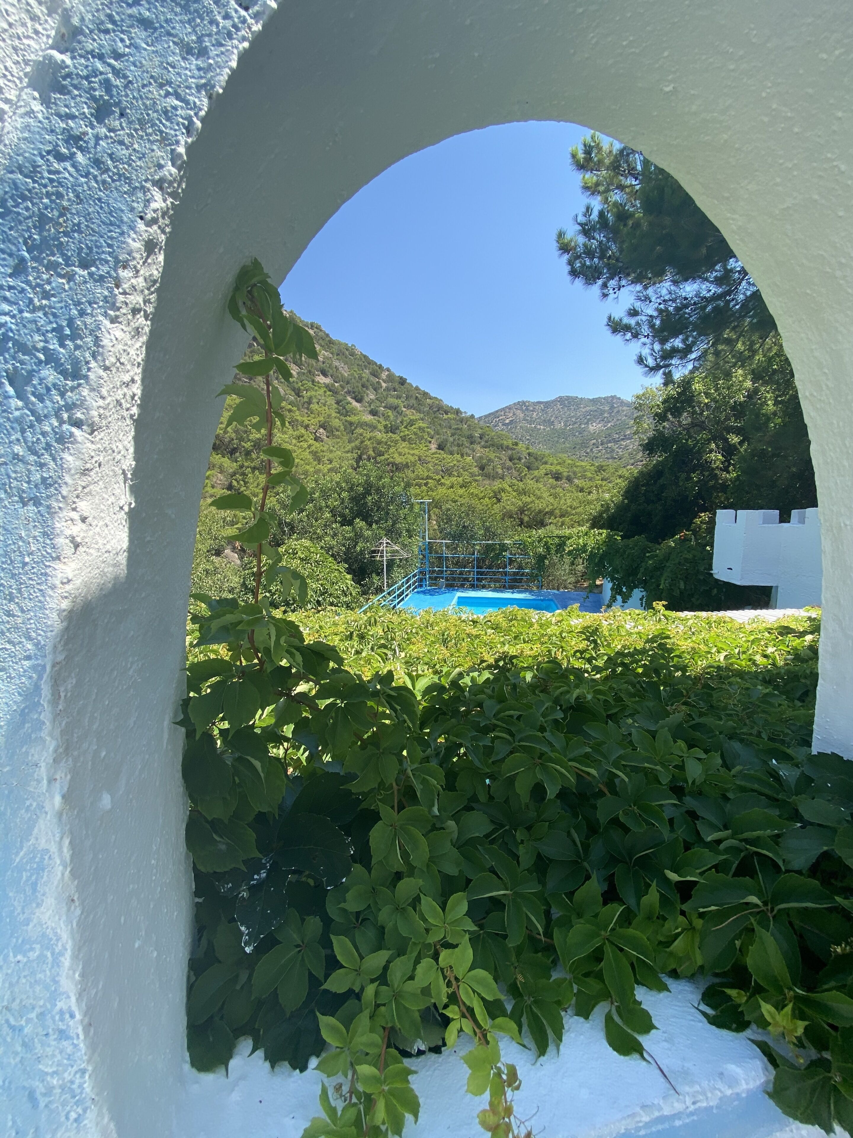 A view through an arched doorway of a pool area surrounded by lush greenery and a mountainous backdrop.