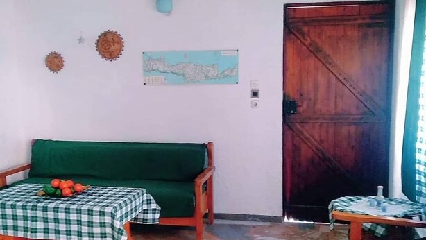 A room with a green sofa, a small table with a checkered tablecloth, a map on the wall, and a wooden door. - Oleander's Garden (Ierapetra)