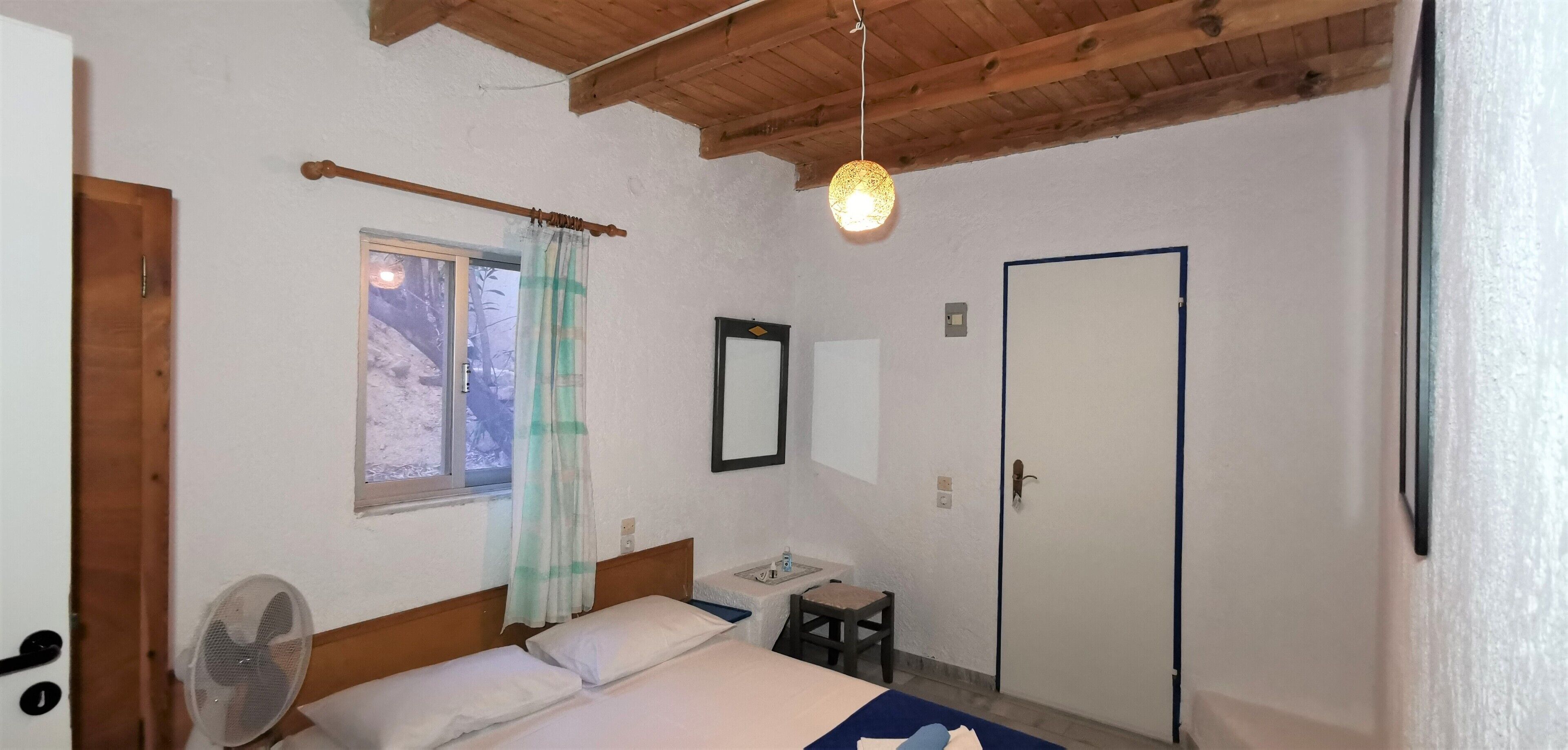Un dormitorio con techo de madera, una cama con ropa de cama blanca, una mesita de noche, un espejo, una ventana con cortina y una puerta.