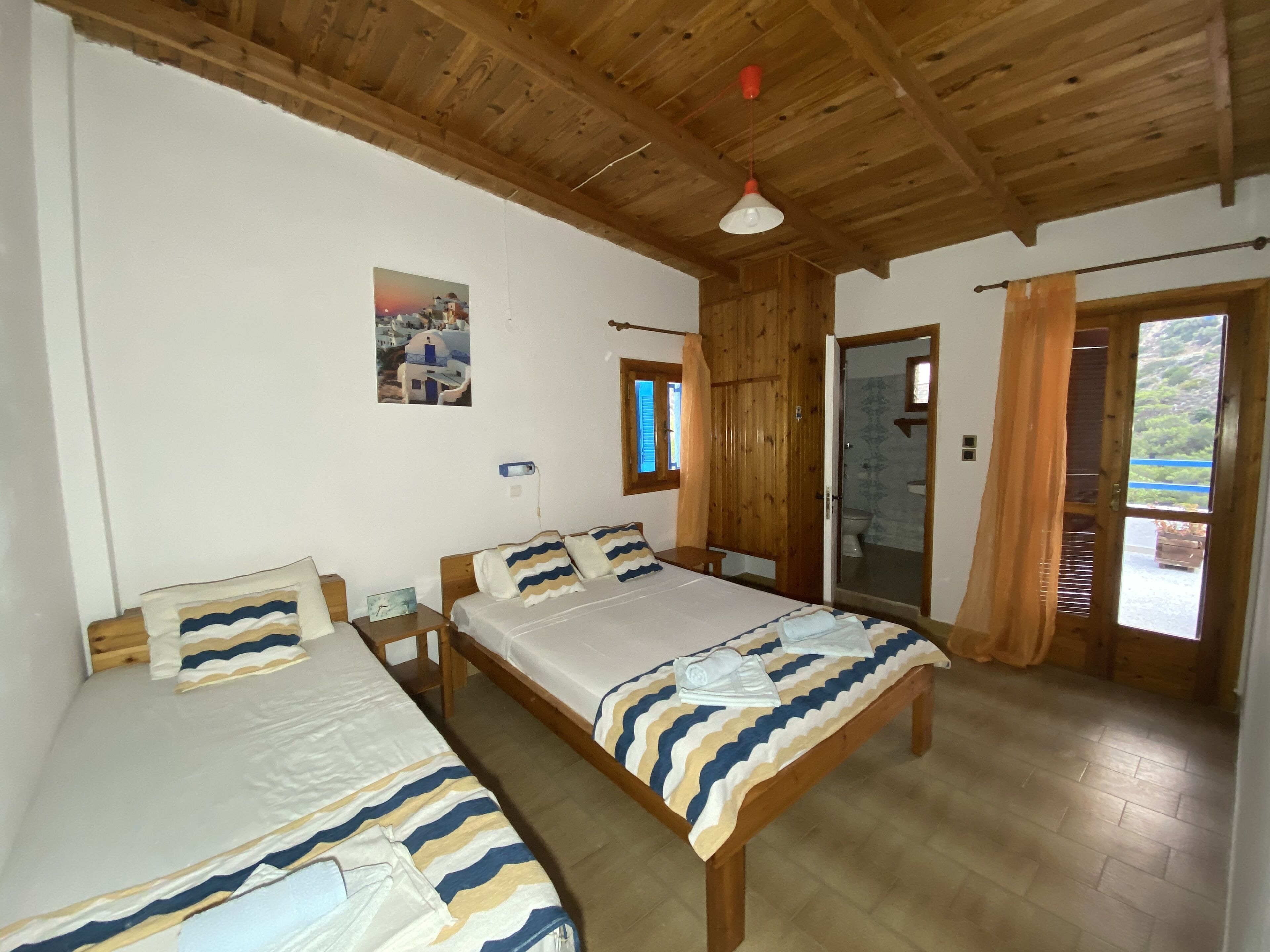 A room with two beds, wooden flooring, a wooden ceiling, a window with curtains, and a door leading to a bathroom.