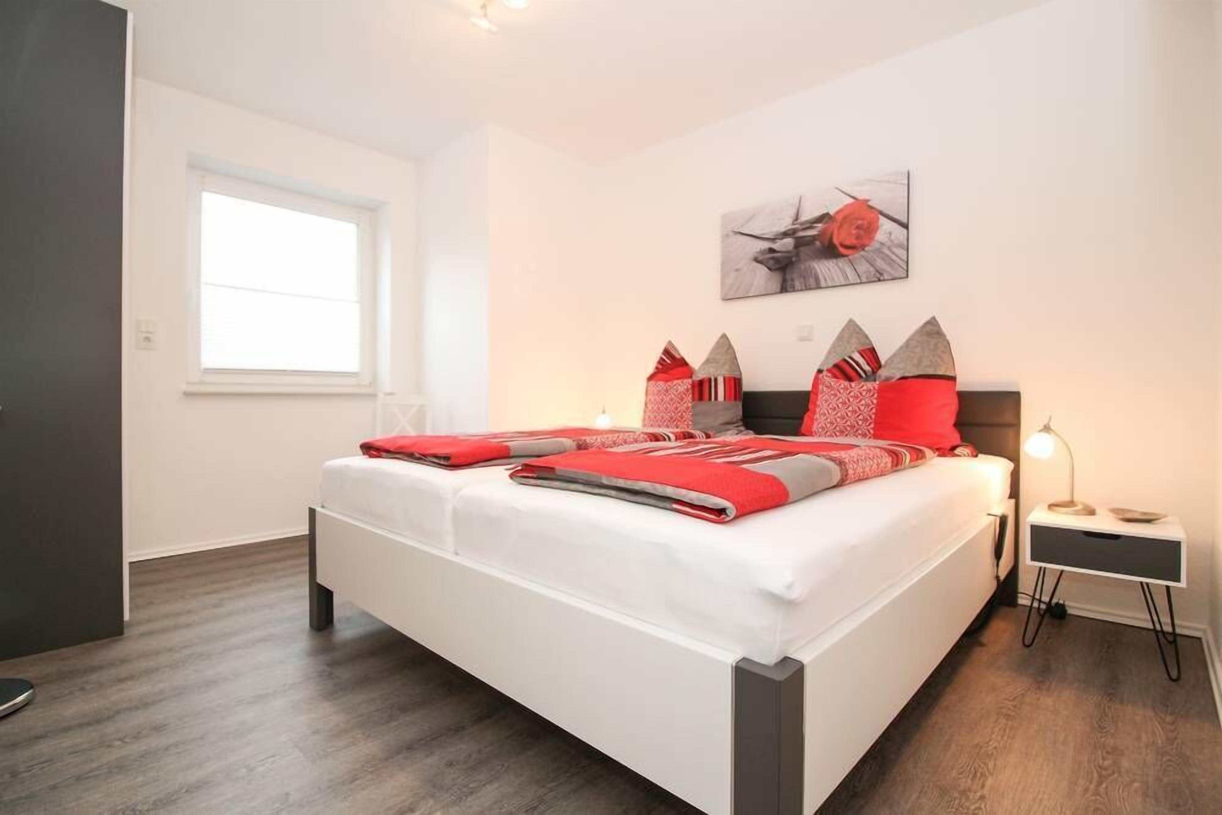A modern bedroom with a large bed, a bedside table with a lamp, a window, and a wall-mounted artwork.