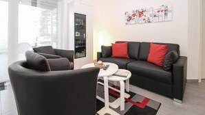Una sala de estar moderna con un sofá de cuero negro, un sillón negro y una mesa de centro blanca. Se observa una estantería con puertas de cristal y un cuadro en la pared.