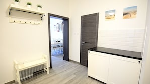 A modern kitchen with white cabinets, a black countertop, and a wooden door. There is a bench with a shelf above it holding potted plants. - Victus Apartamenty - Riviera (Sopot)