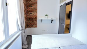 A small, well-lit room with a white bed, a brick wall, a small shelf with a plant, and a door leading to another room. - Victus Apartamenty - Riviera (Sopot)