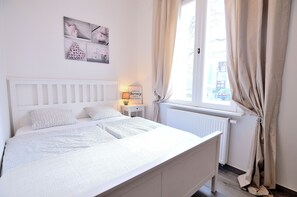 Cama bem arrumada com cabeceira branca, mesa de cabeceira com abajur e janela com cortinas.