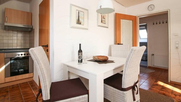 A modern kitchen with white cabinetry, a built-in oven, and a dining area with a white table and wicker chairs. - Muckelstuuv (Krummhoern)