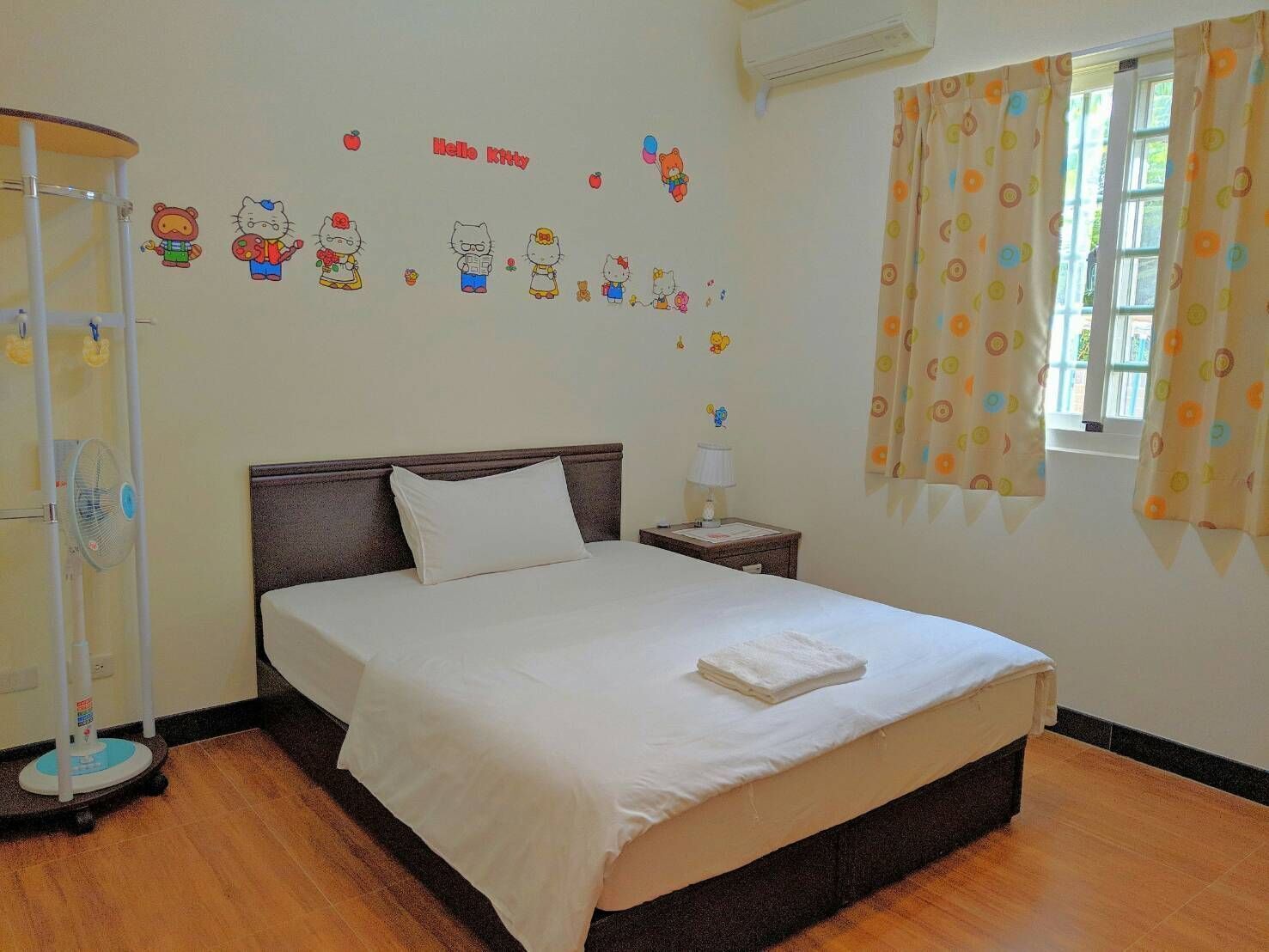 Una cama bien hecha con edredón blanco y almohadas, una mesita de noche con una lámpara, una pared con adornos de Hello Kitty y una ventana con cortinas.