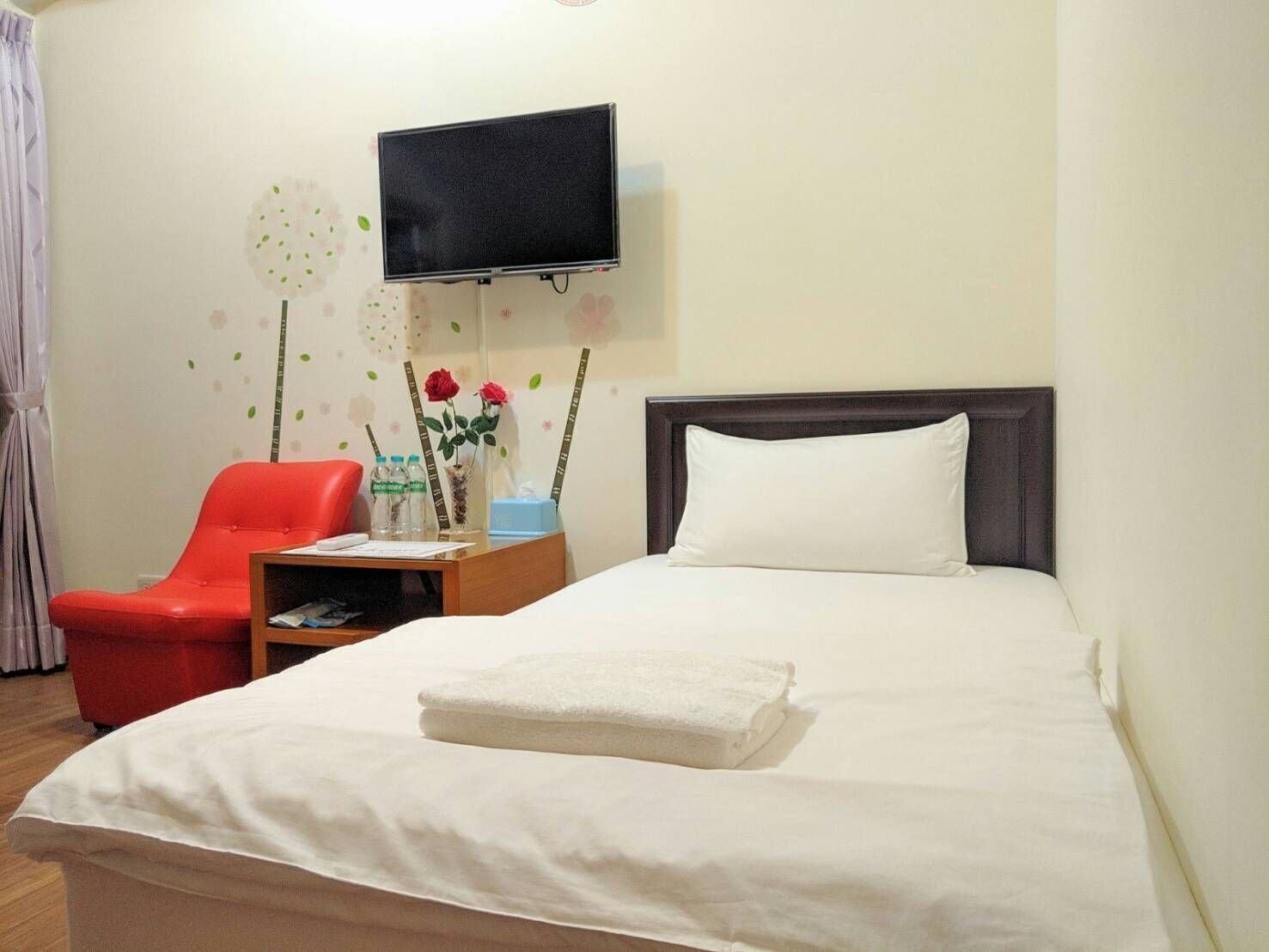 Una habitación de hotel con una cama, un televisor montado en la pared, una silla roja y una mesita de noche con un florero.