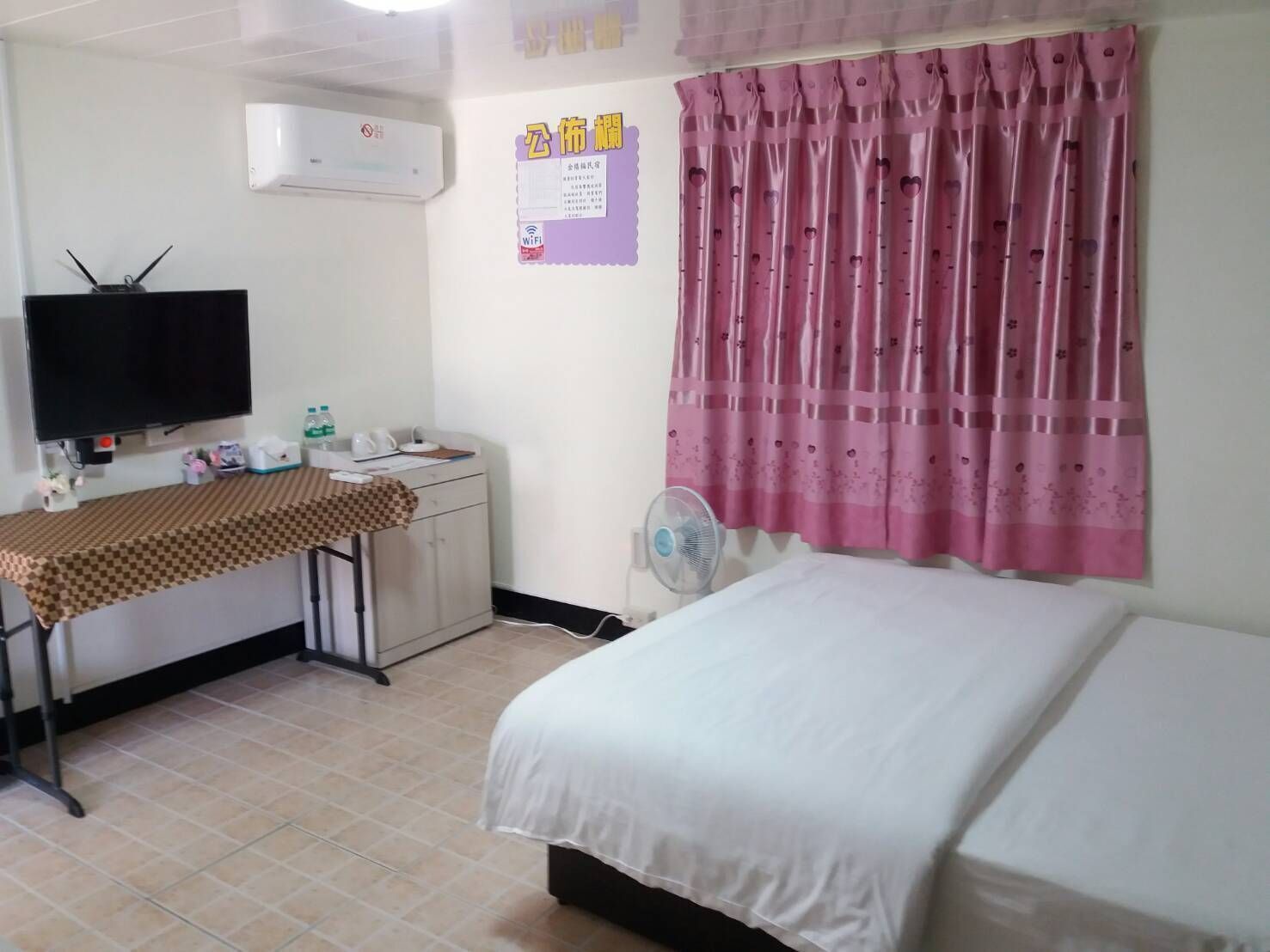 Een hotelkamer met een bed, een bureau met een stoel, een televisietoestel, een airconditioner, een ventilator en een raam met roze gordijnen.