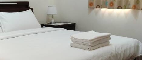 Una cama bien tendida con sábanas blancas, cabecera de madera, mesita de noche con lámpara y ventana con cortinas estampadas.