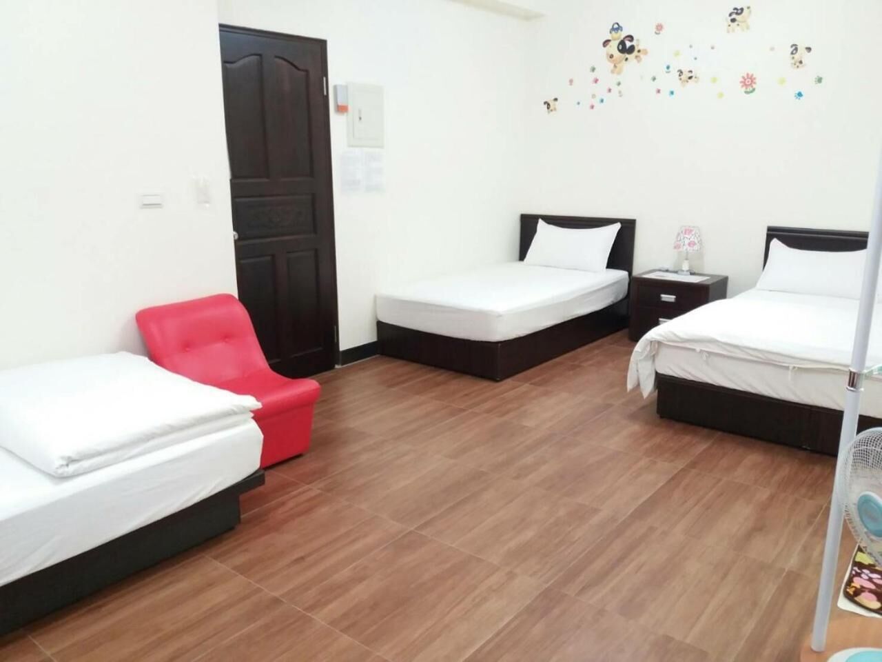 A hotel room with two single beds, a red chair, a small table, and a wall-mounted fan.