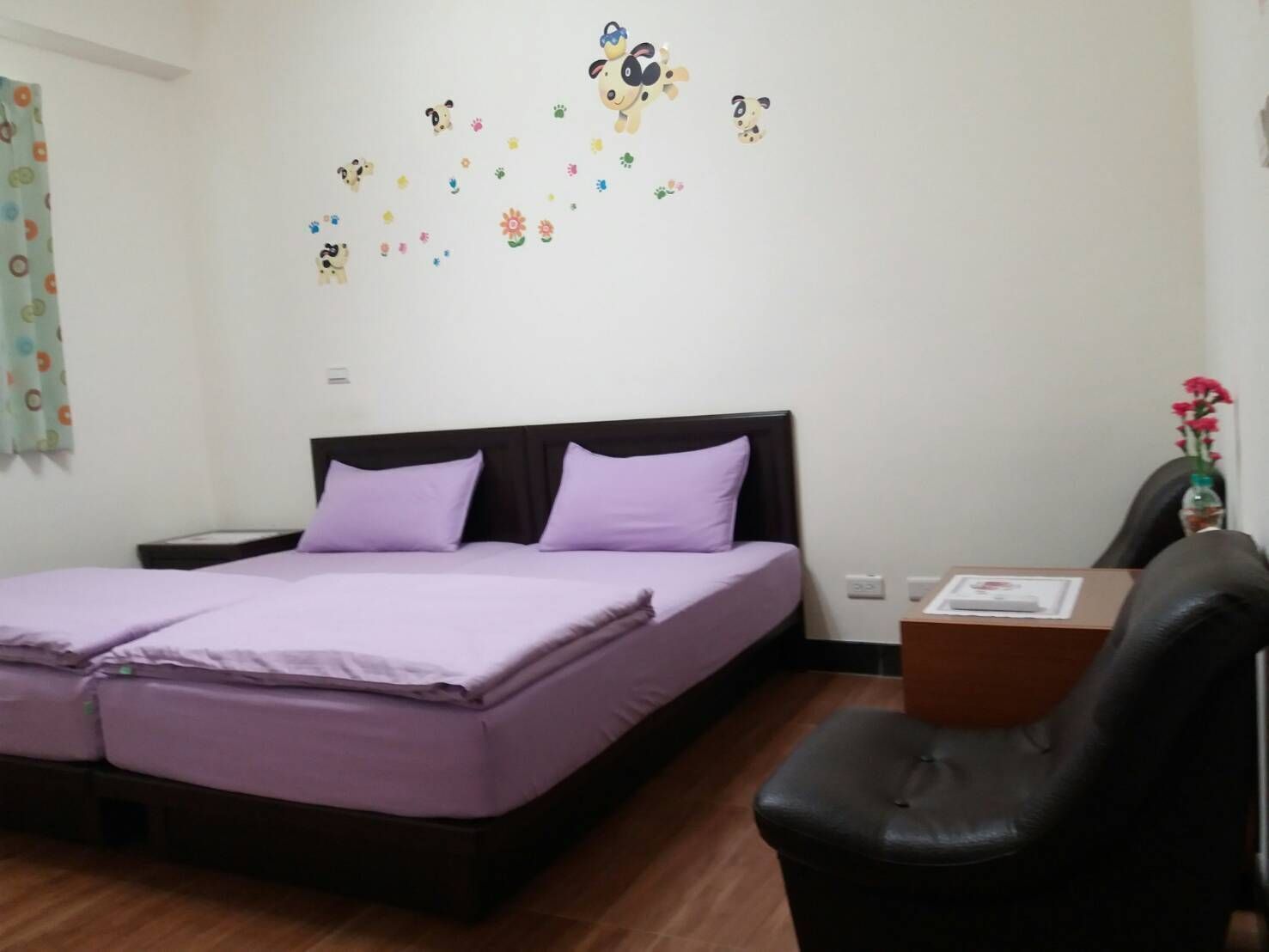 Habitación de hotel con cama doble, dos almohadas, una mesita de noche, un escritorio con una silla y un adhesivo mural de animales y flores.