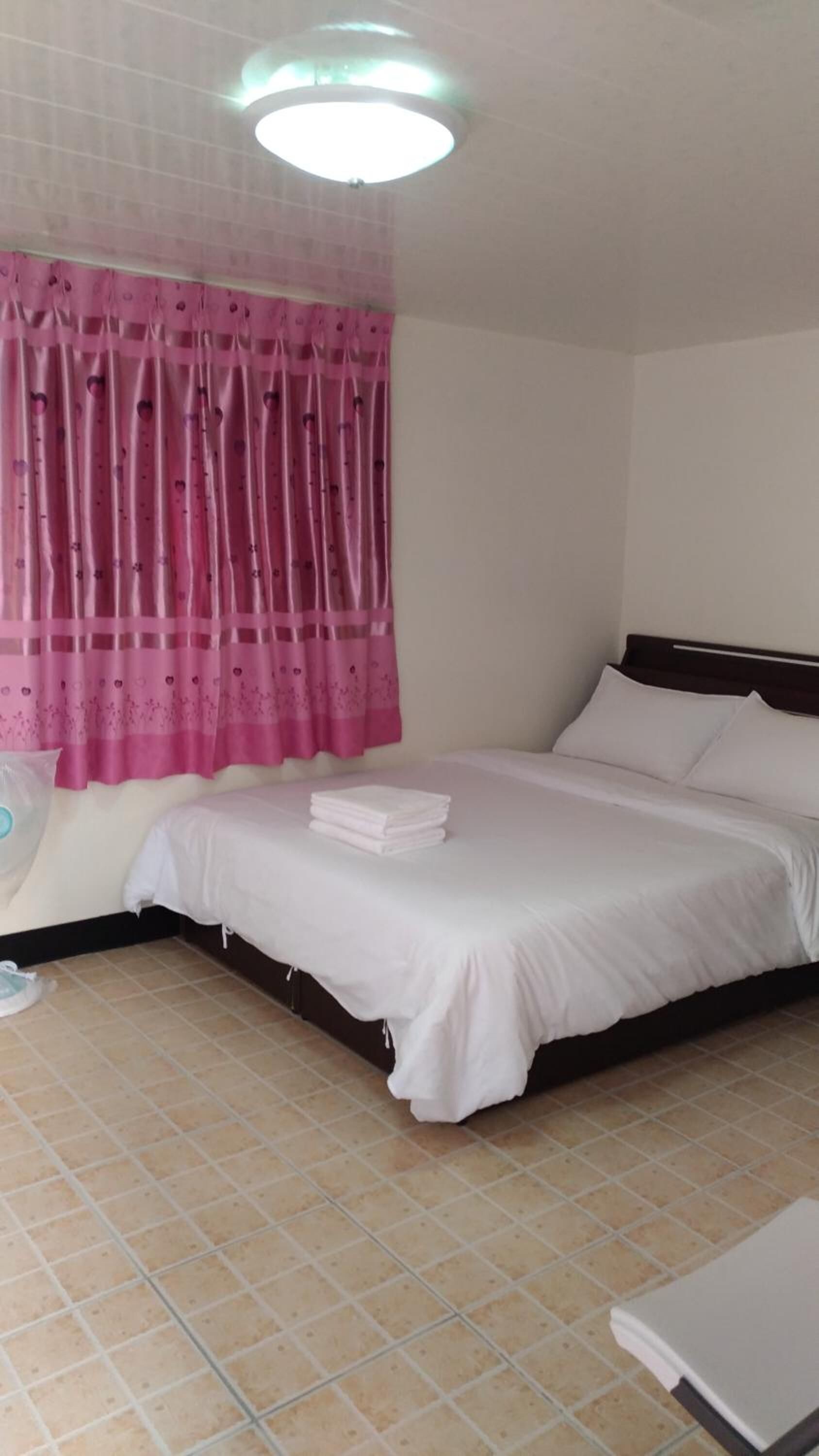 A bedroom with a bed, pillows, a bedside table, a wall-mounted fan, and a window with pink curtains.