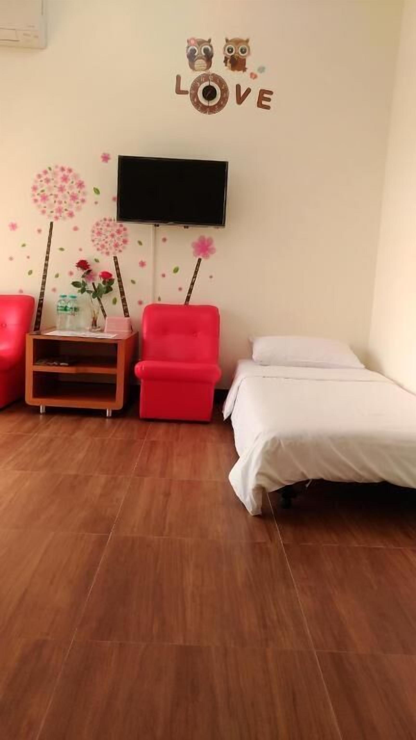 Una habitación con una cama, una silla roja, una pequeña mesa de madera con un jarrón de flores y un televisor montado en la pared.