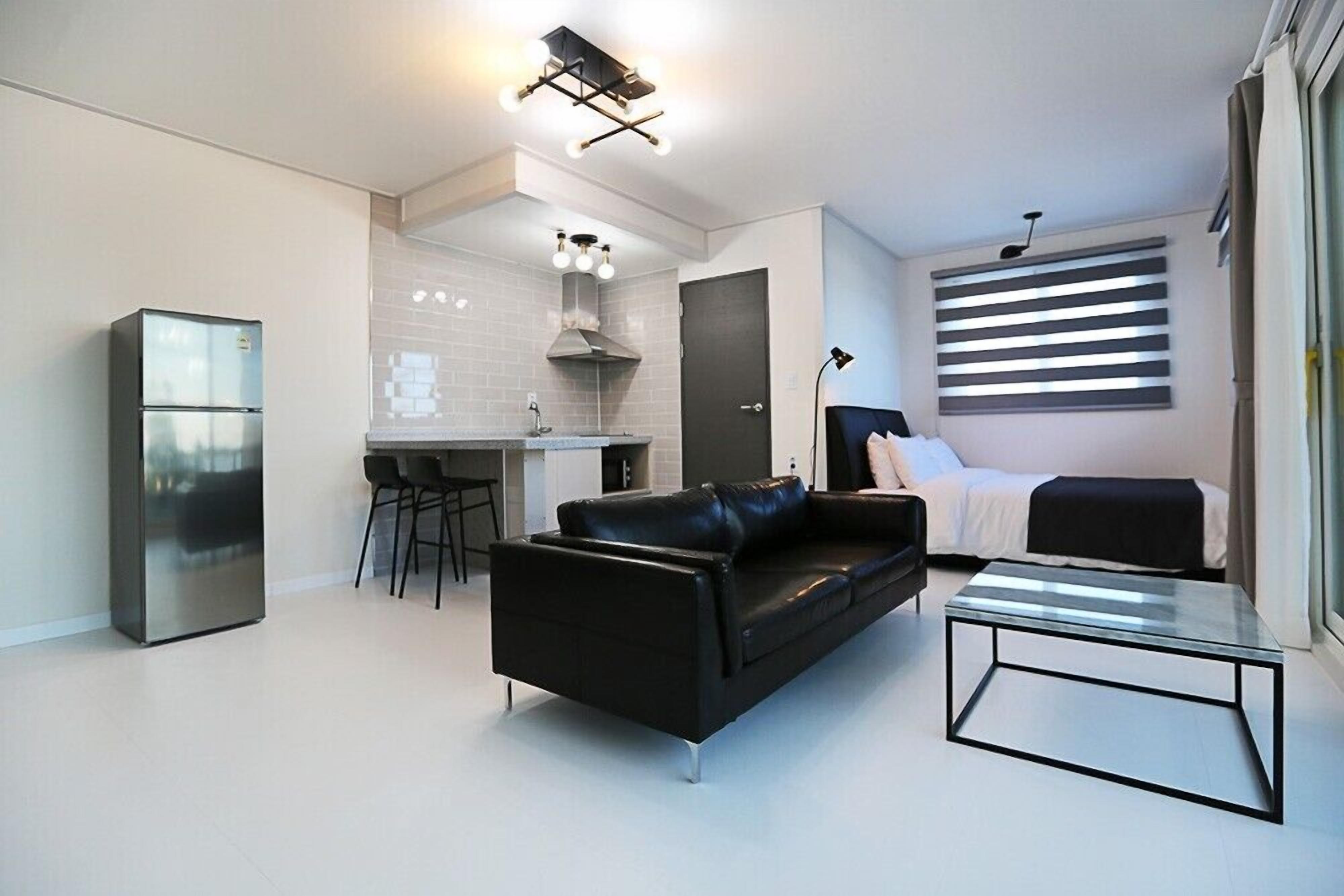 Un espace de vie moderne comprenant un canapé en cuir noir, une table basse en verre, un coin cuisine avec un réfrigérateur et un ensemble de salle à manger.