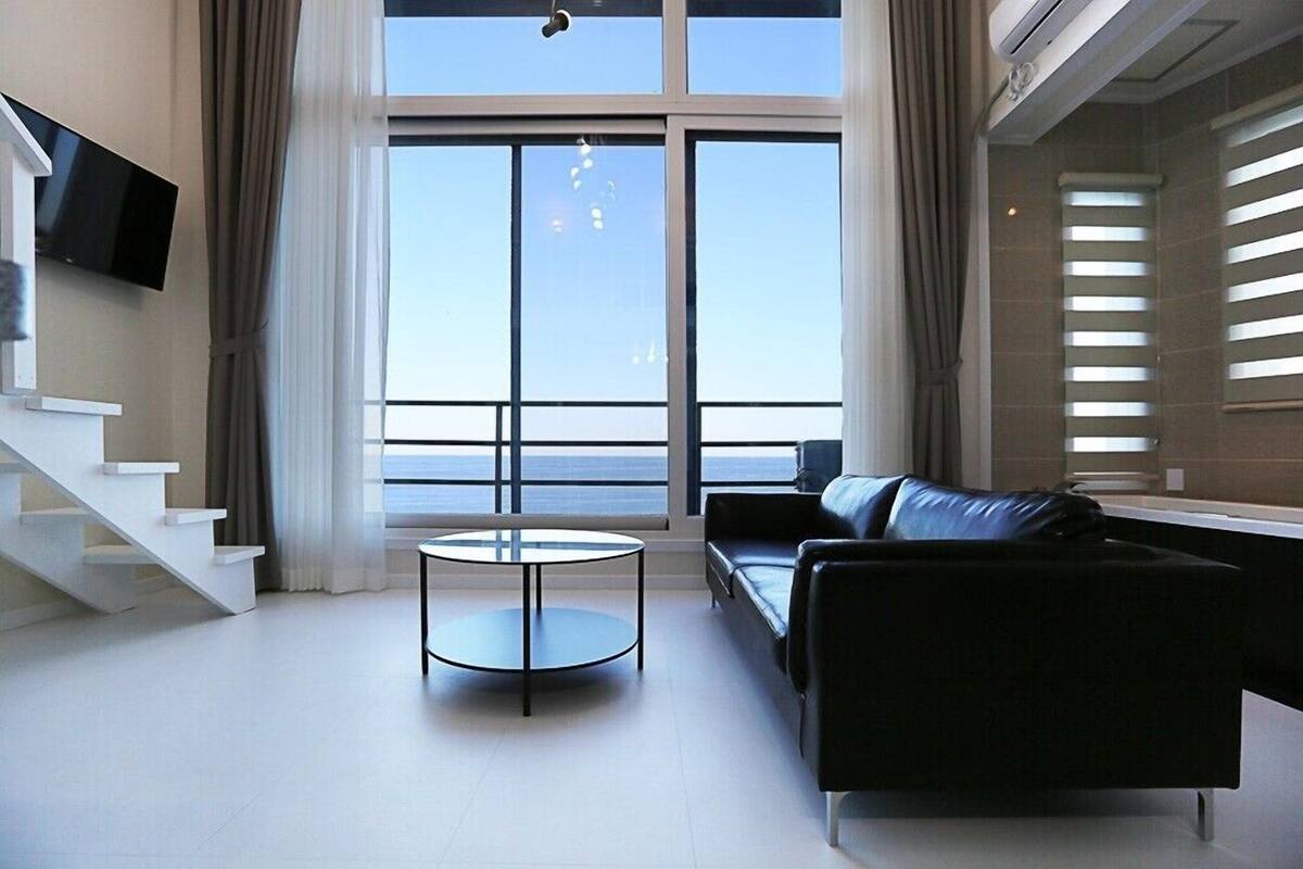 A modern living room with a large window, a black leather sofa, a small round table, and a staircase.