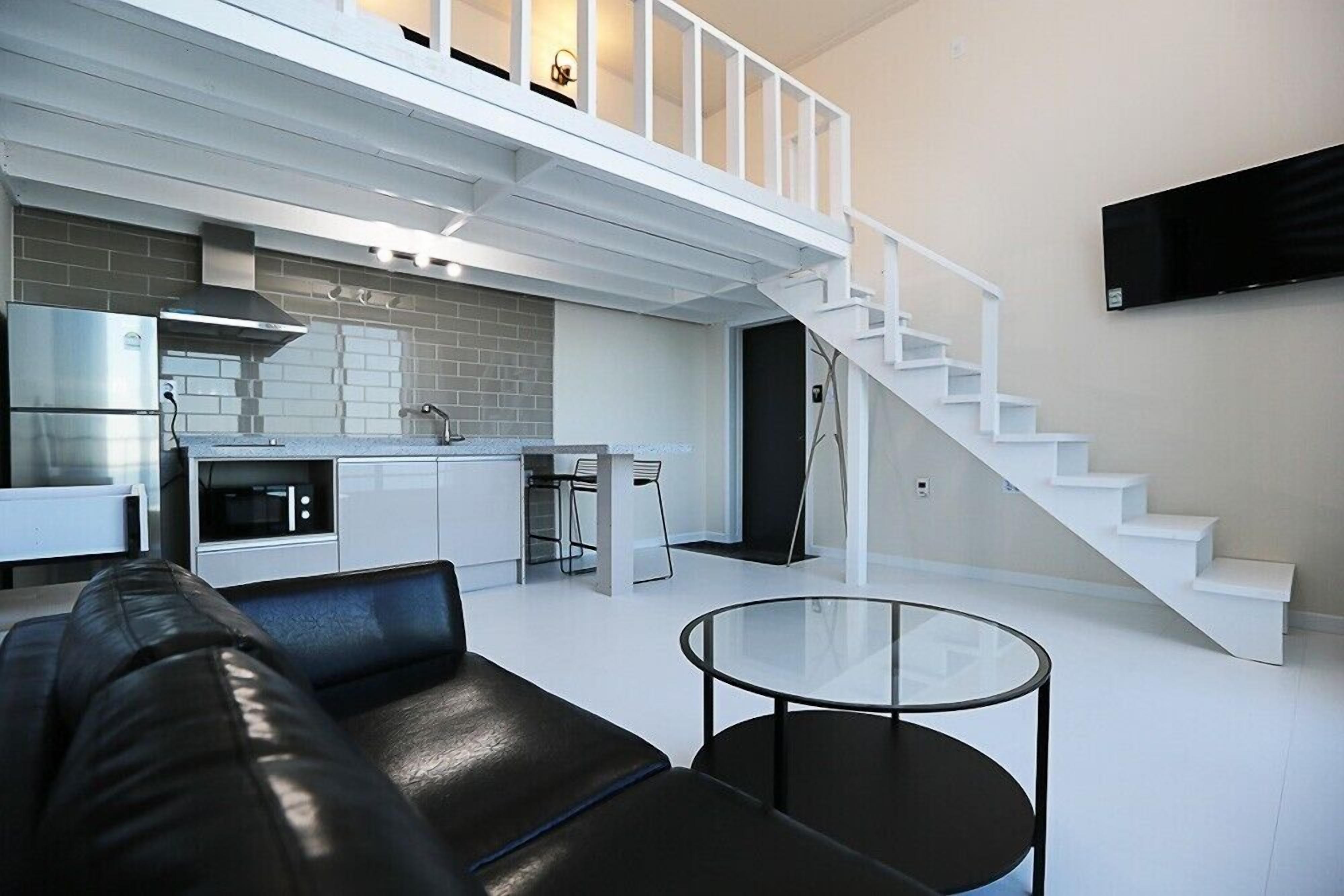 Un espacio moderno para vivir con un sofá de cuero negro, una mesa de centro de vidrio, una zona de cocina con una escalera blanca y un televisor de pantalla plana montado en la pared.