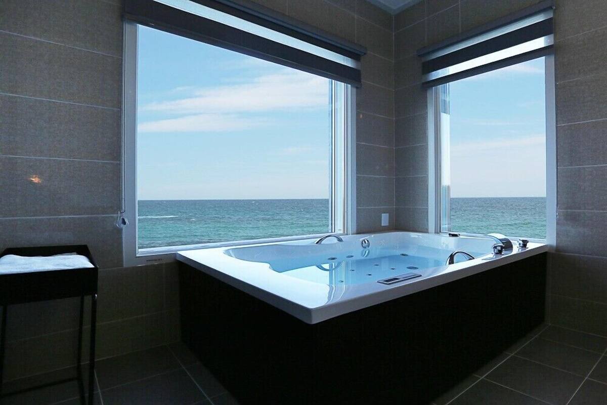 A modern bathroom with a large bathtub and a view of the sea through two windows.