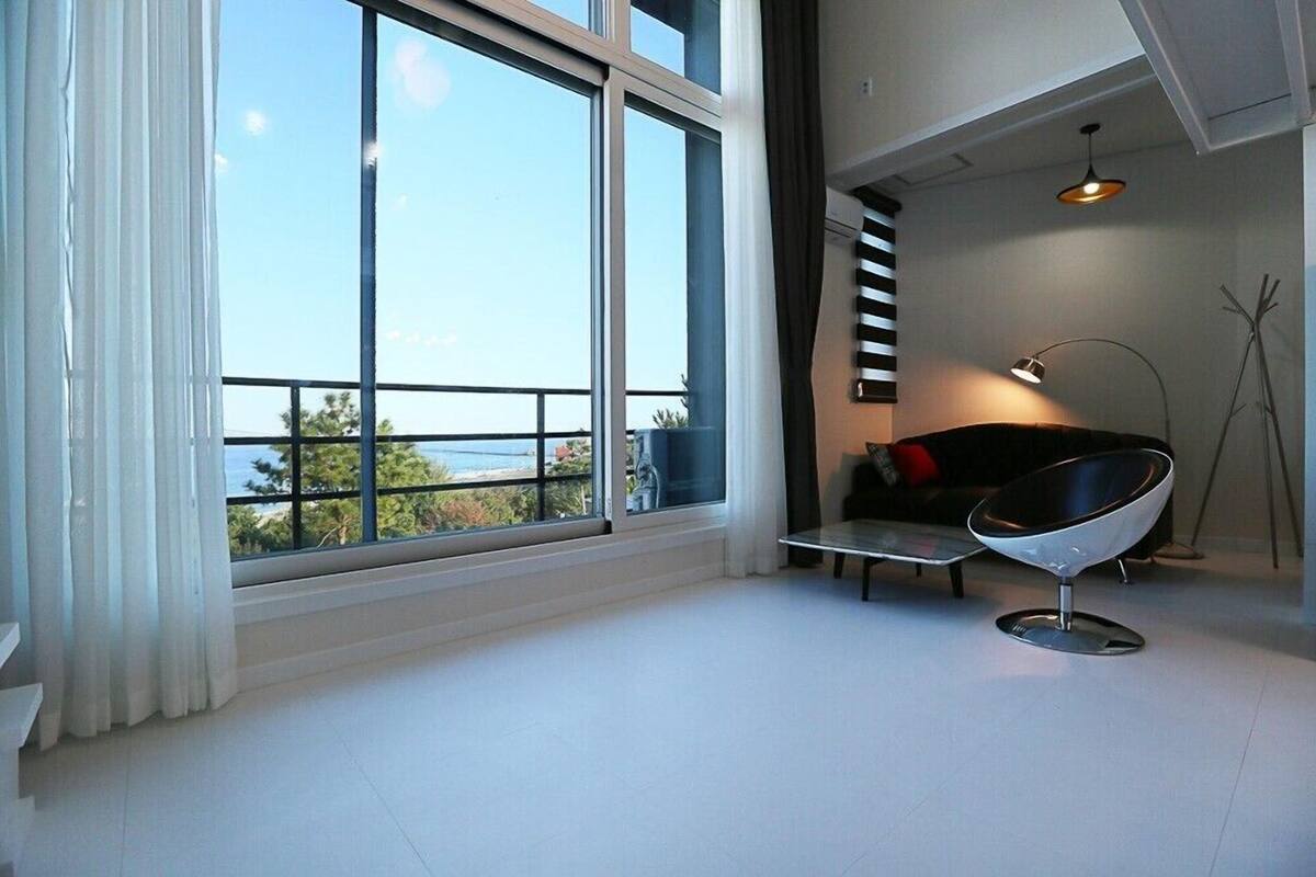 A modern living room with large windows, a black chair, a small table, and a sofa with a red cushion.