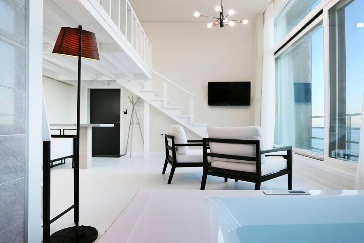 A modern hotel room with a large window, a black chair, a floor lamp, and a flat-screen TV mounted on the wall.