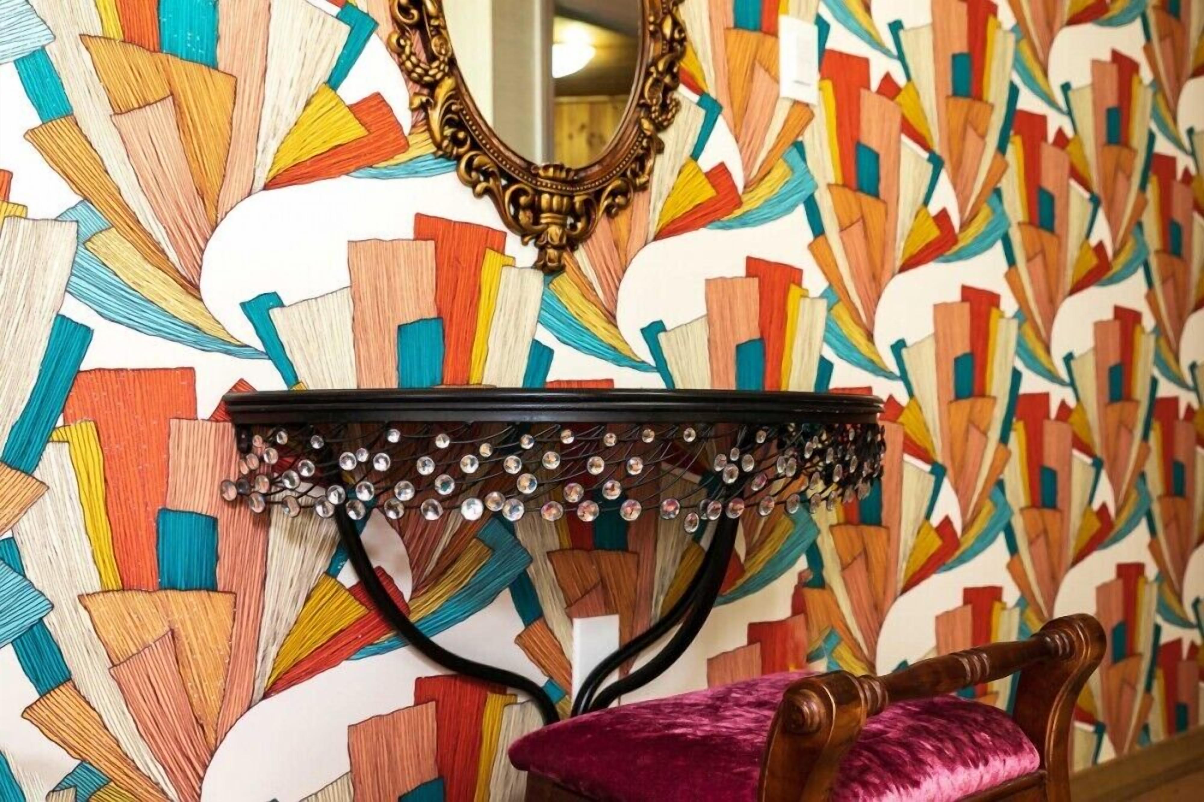 Una habitación con una pared de patrón geométrico y coloridos motivos, una silla de madera con cojín rosa y un espejo decorativo sobre una consola cubierta de espejos.