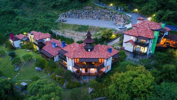 A multi-story building with a red-tiled roof, surrounded by lush greenery and a stone pathway. - Yangpyung Lavin Pension (Yangpyeong)