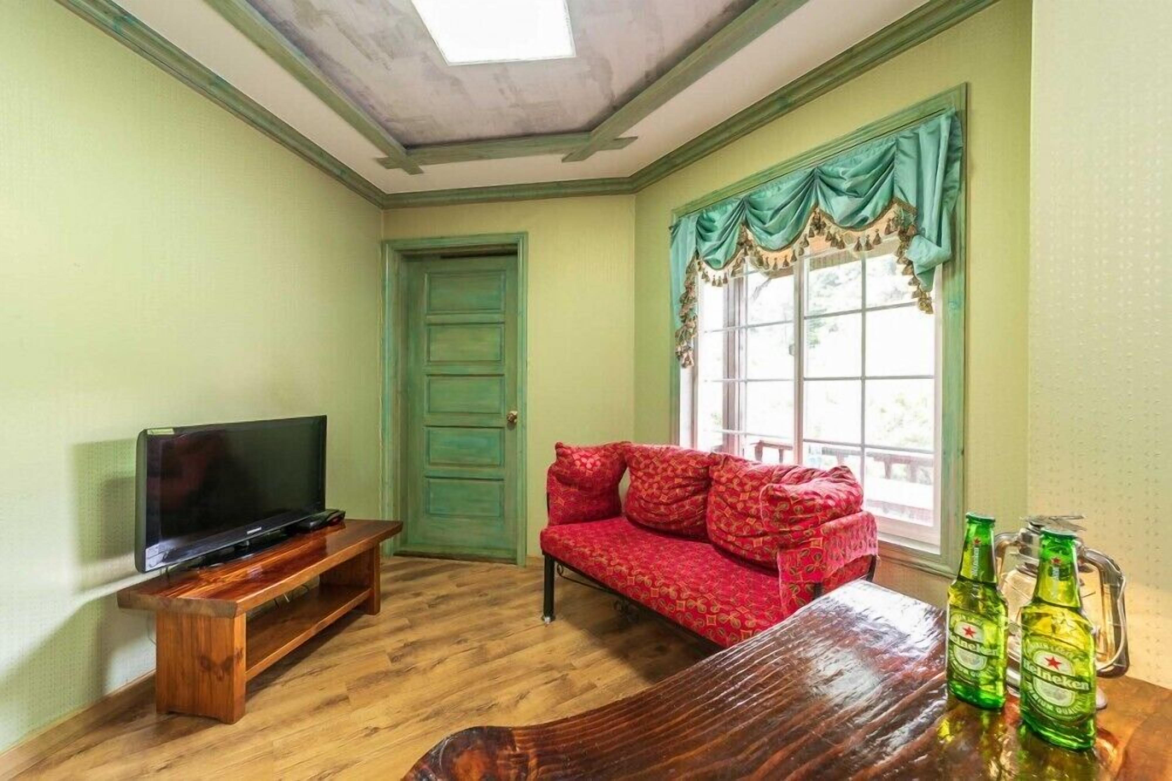 A room with a wooden TV stand, a red sofa, a dining table with beer bottles, and a window with green curtains.