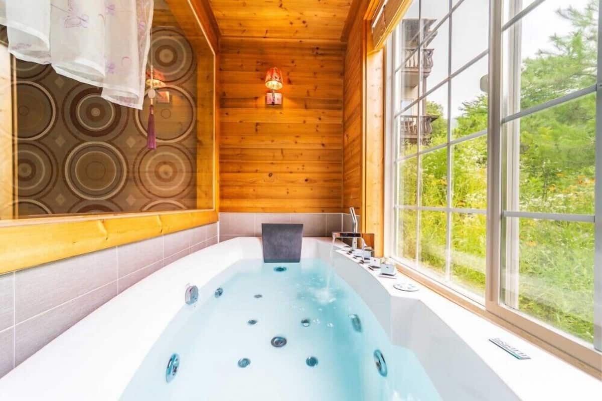 A modern bathroom with a large bathtub, wooden ceiling, and a view of greenery outside.